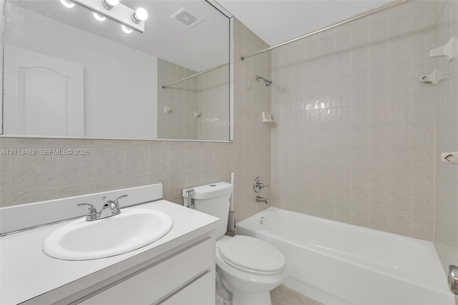 Bathroom featuring a vanity with a white countertop and oval under-mount sink, large wall-mounted mirror, overhead vanity lighting, and a shower-tub combination with ceramic tile surround