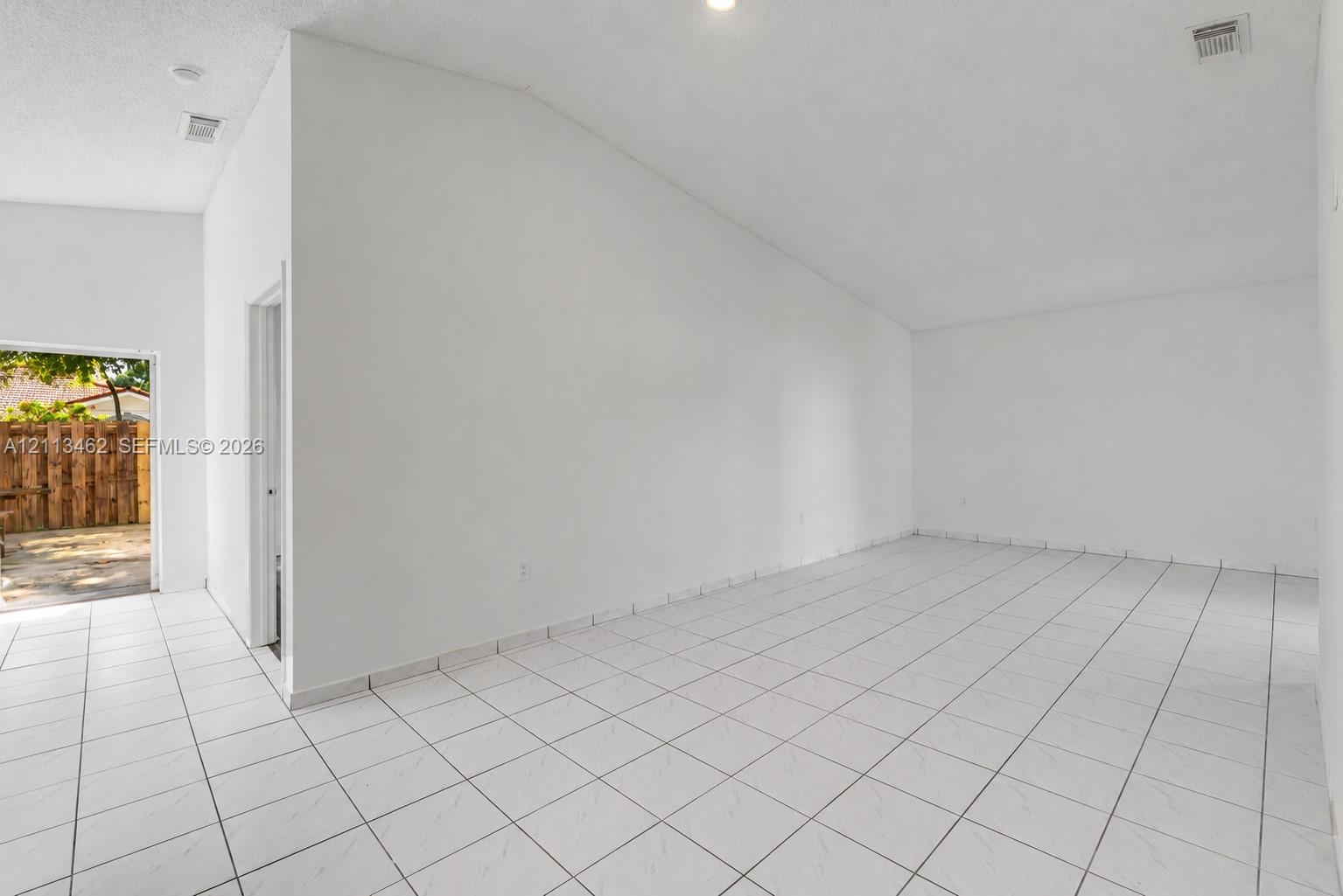 High-ceiling interior space featuring large-format ceramic tile flooring, neutral wall finishes, and an exterior doorway with access to a paved patio area