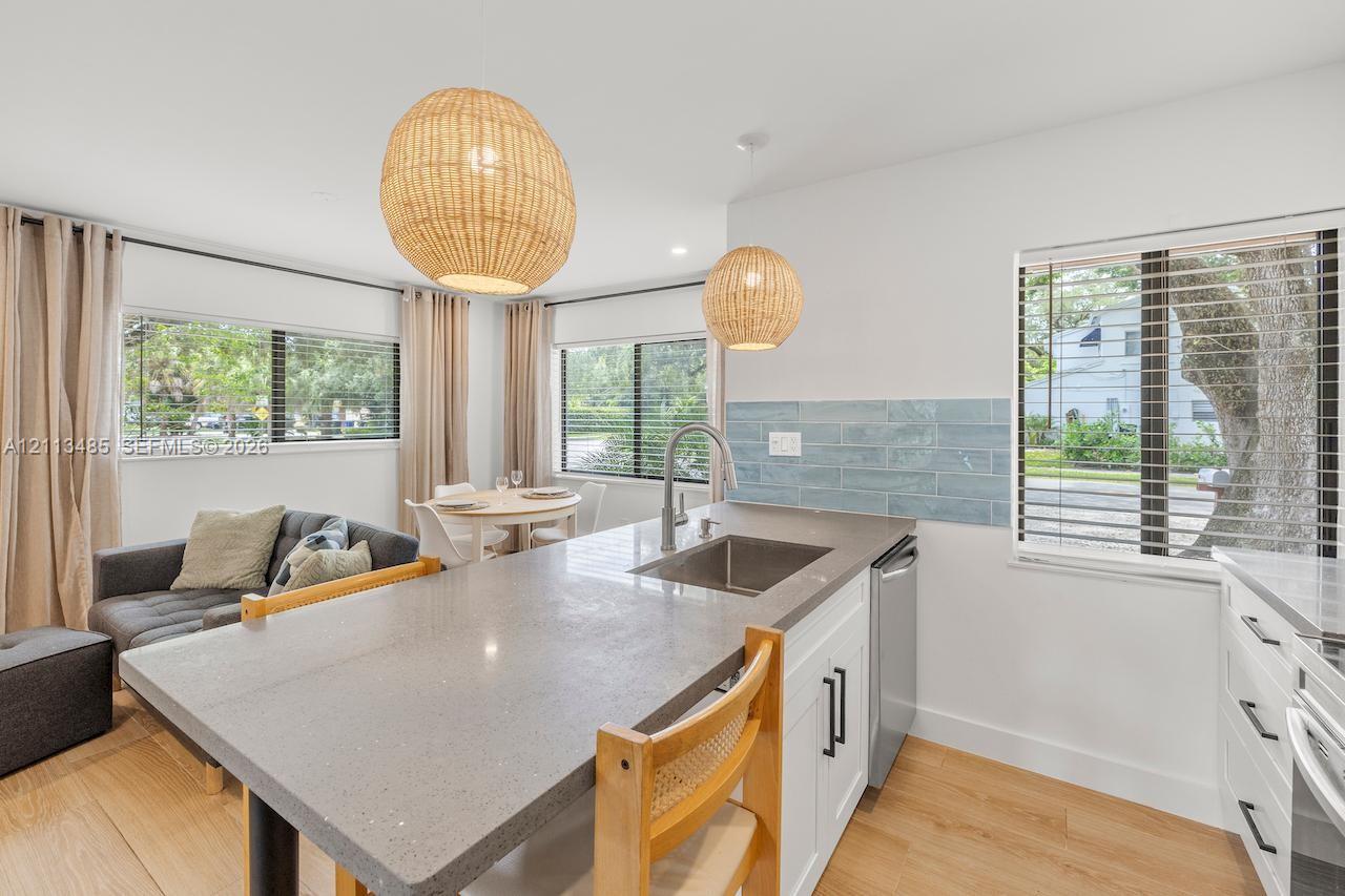 Kitchen featuring a gray composite countertop island, stainless steel undermount sink, blue subway tile backsplash, white cabinetry with matte black hardware, and woven pendant light fixtures