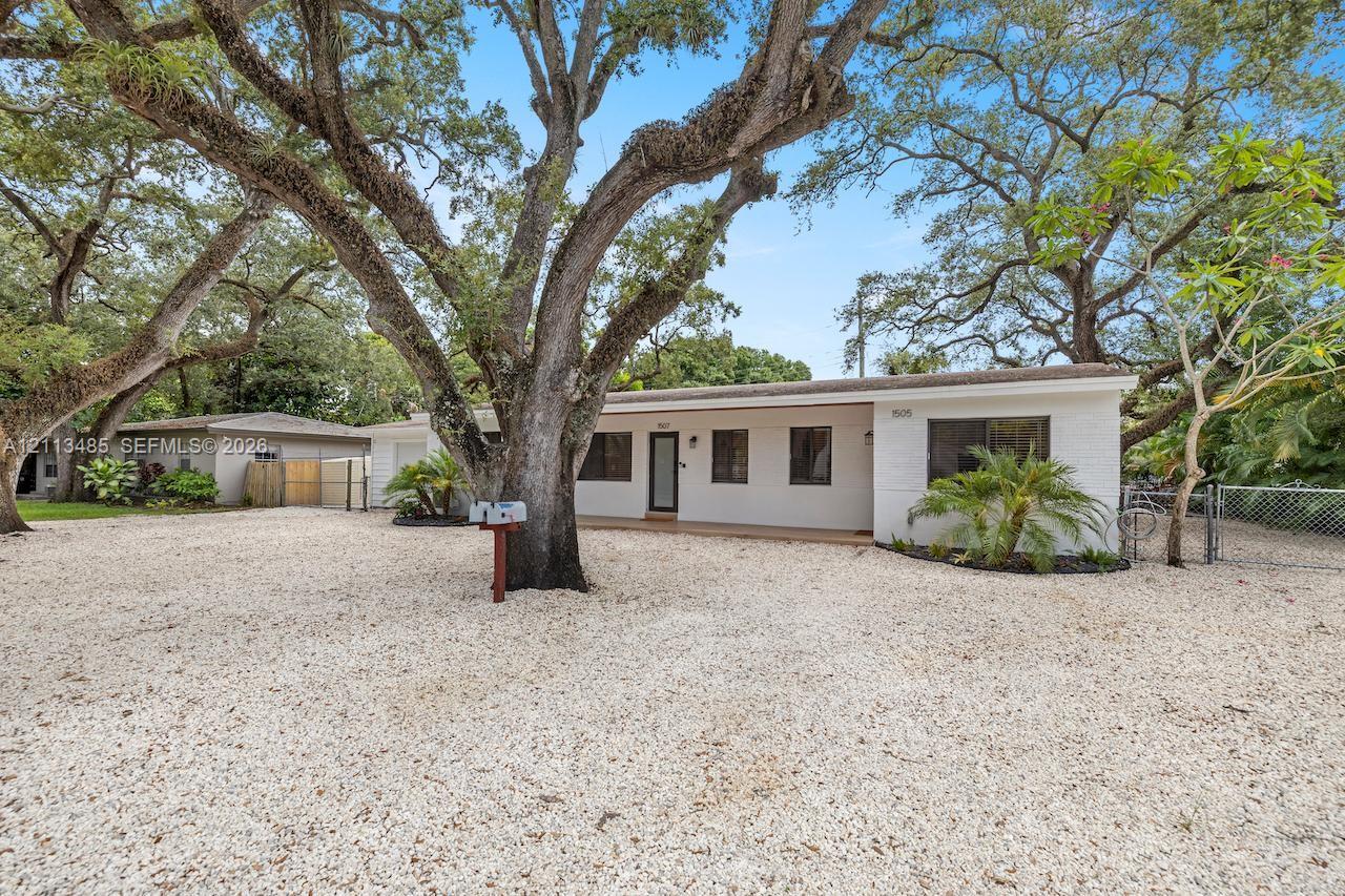 Single-story residence featuring a painted masonry exterior, extensive gravel surfacing, mature oak trees, and chain-link fencing
