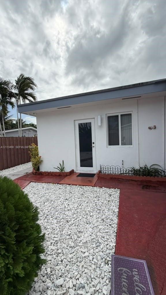 Exterior featuring white stucco siding, a glass-panel entry door, a red painted concrete patio, and white decorative stone ground cover