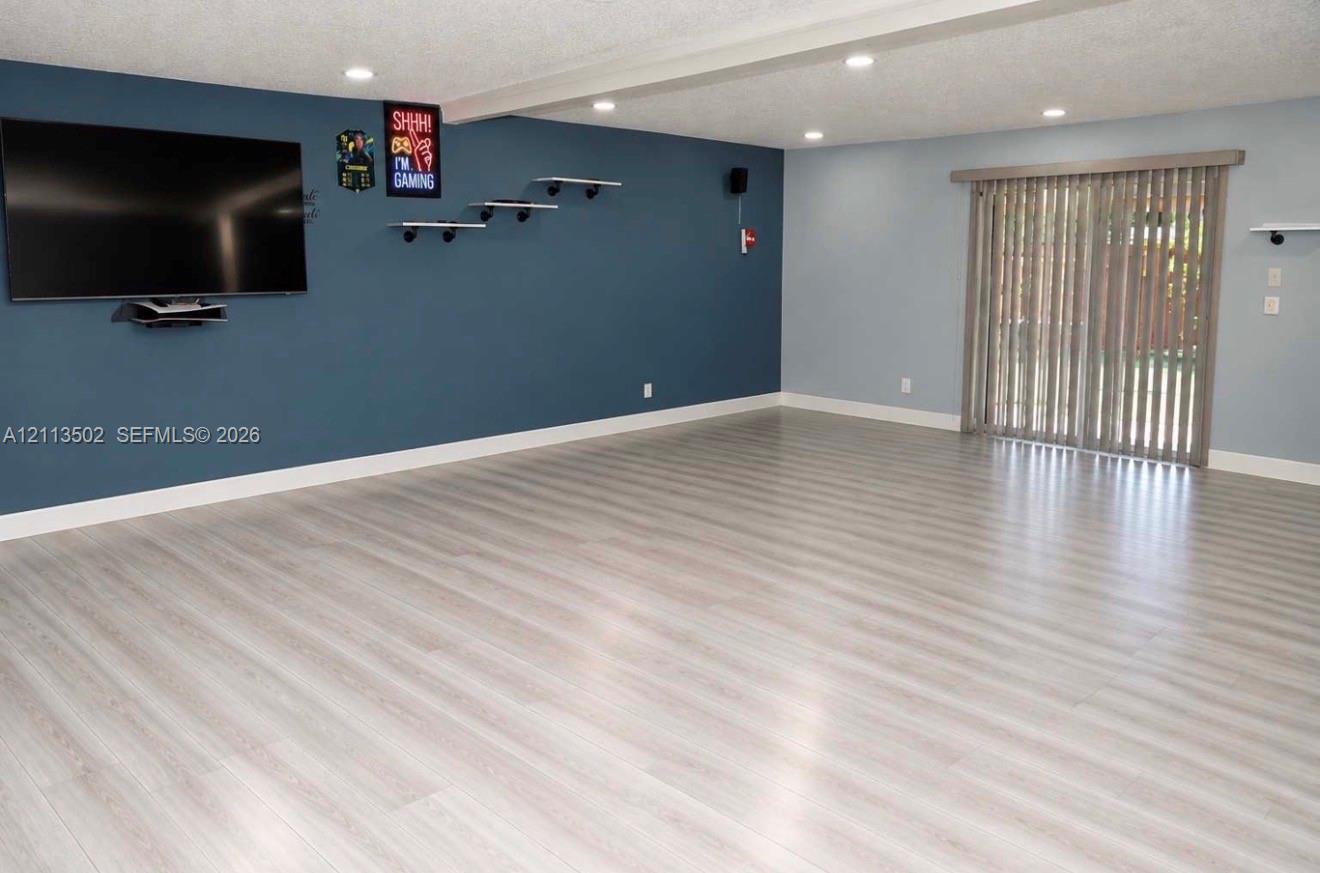 Interior space featuring wood-finish flooring, recessed lighting, a dark-toned accent wall with wall-mounted shelving, and sliding glass doors with vertical blinds Interior space featuring wood-finish flooring, recessed lighting, a dark-toned accent wall with wall-mounted shelving, and sliding glass doors with vertical blinds