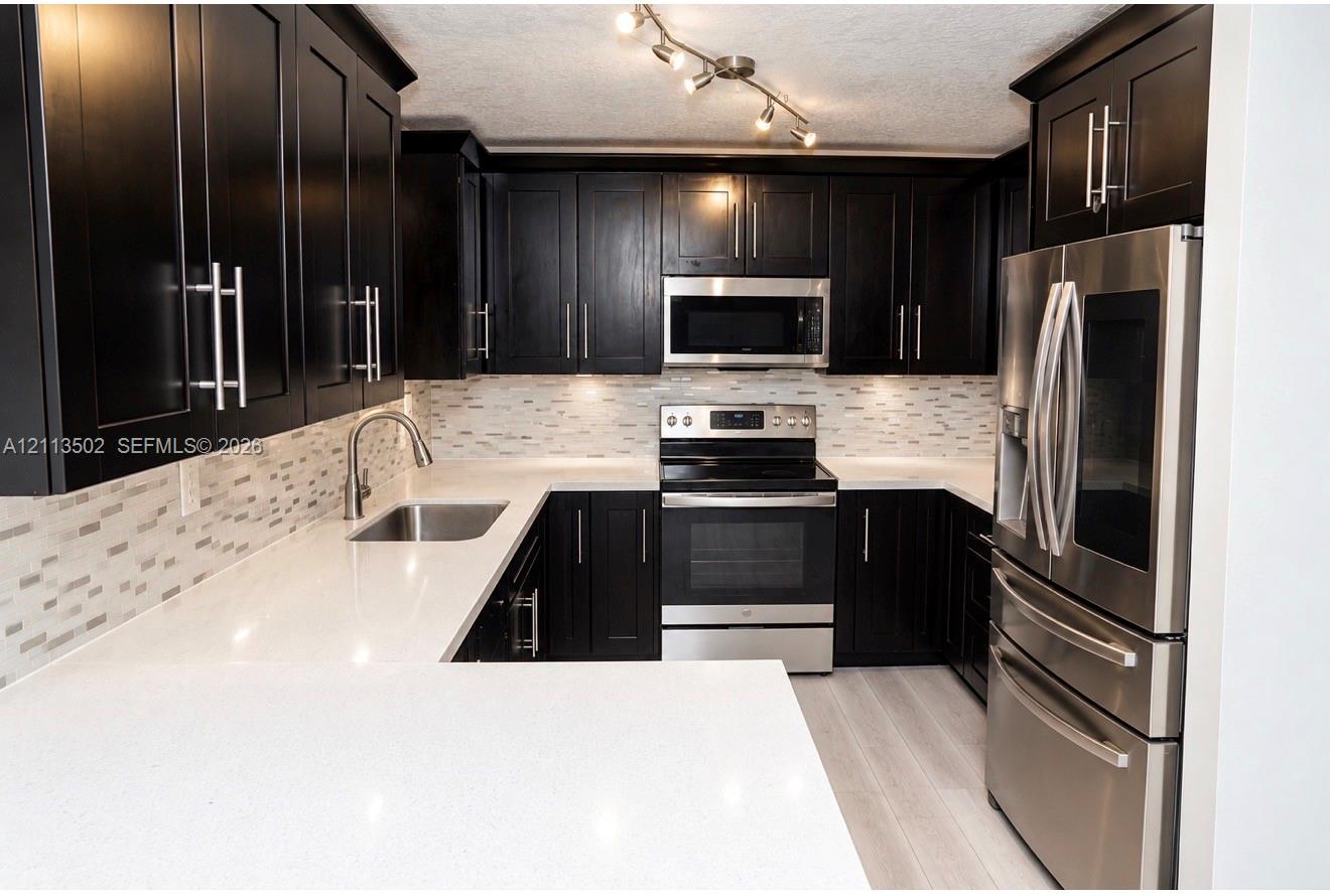 Kitchen featuring dark shaker-style cabinetry, white quartz countertops, multi-toned linear tile backsplash, stainless steel appliances, and track lighting Kitchen featuring dark shaker-style cabinetry, white quartz countertops, multi-toned linear tile backsplash, stainless steel appliances, and track lighting