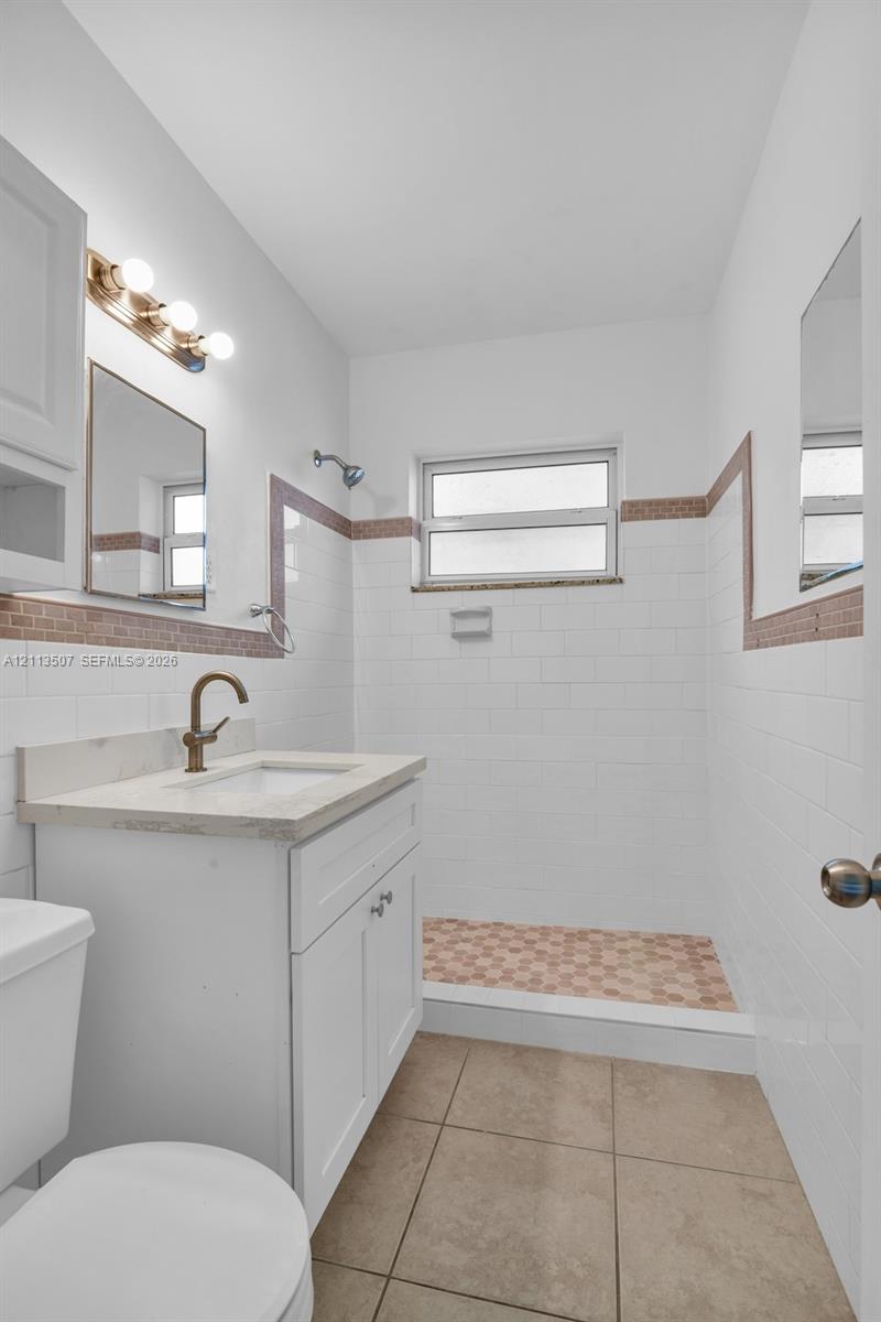 Bathroom featuring a white vanity with a neutral stone countertop, bronze-finish faucet, white subway tile walls with decorative border accents, walk-in shower with mosaic tile flooring, and large-format tile flooring Bathroom featuring a white vanity with a neutral stone countertop, bronze-finish faucet, white subway tile walls with decorative border accents, walk-in shower with mosaic tile flooring, and large-format tile flooring
