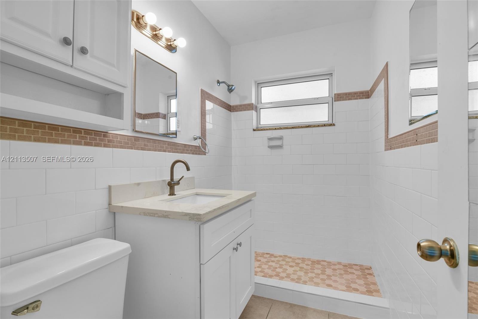 Bathroom featuring white subway tile walls with decorative mosaic inlay, a walk-in shower with mosaic tile flooring, a white vanity with a neutral-toned countertop, and a wall-mounted vanity light fixture Bathroom featuring white subway tile walls with decorative mosaic inlay, a walk-in shower with mosaic tile flooring, a white vanity with a neutral-toned countertop, and a wall-mounted vanity light fixture