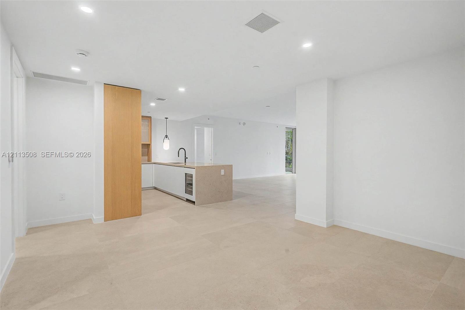 Open-concept interior featuring large-format tile flooring, recessed ceiling lighting, a kitchen island with a stone-finish countertop, integrated under-counter appliance, and wood-finish cabinetry
