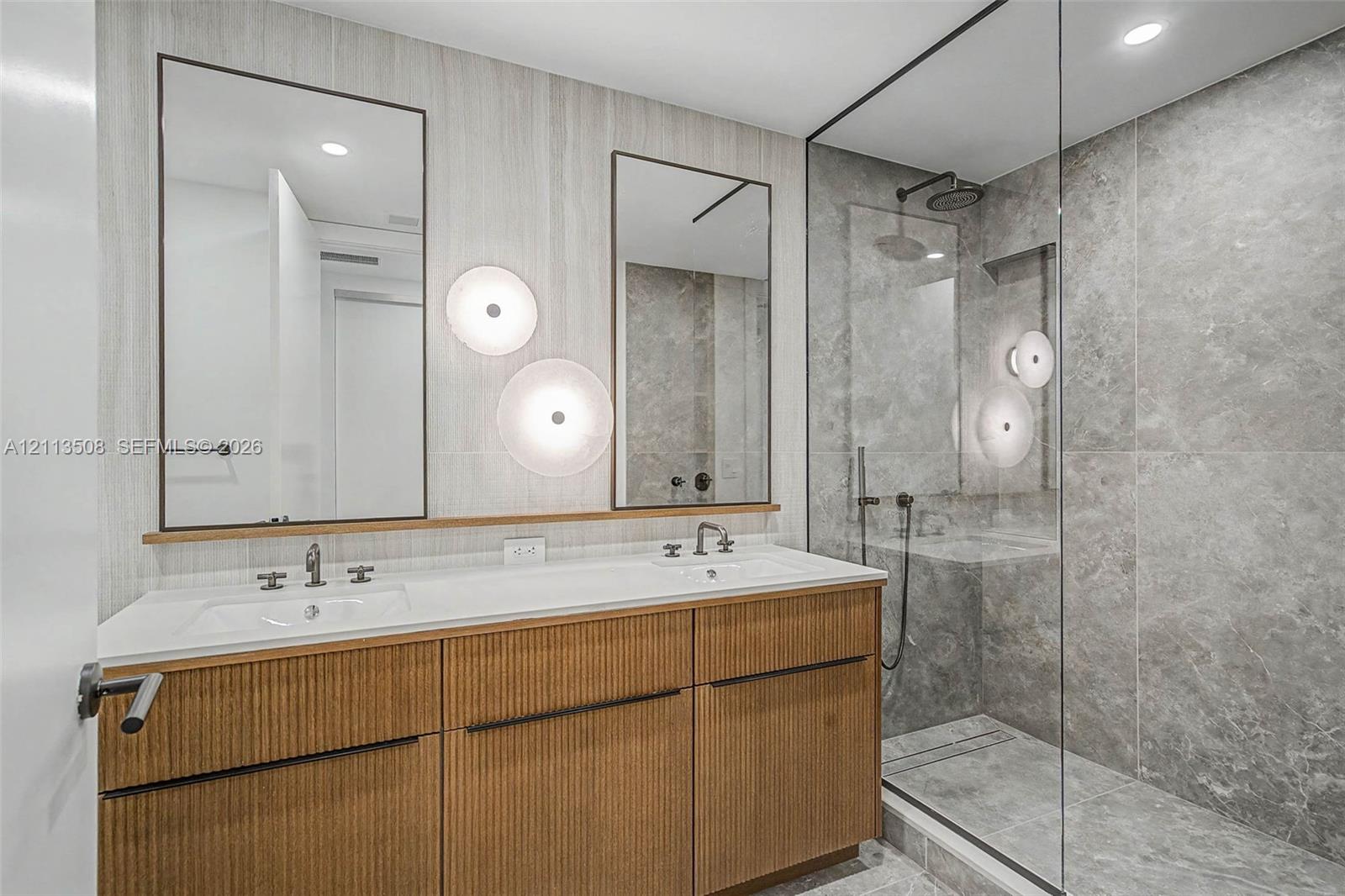 Dual-sink vanity with fluted wood-finish cabinetry, rectangular under-mount basins, wall-mounted round lighting, and a glass-enclosed shower featuring large-format stone-style tile and rain-style showerhead