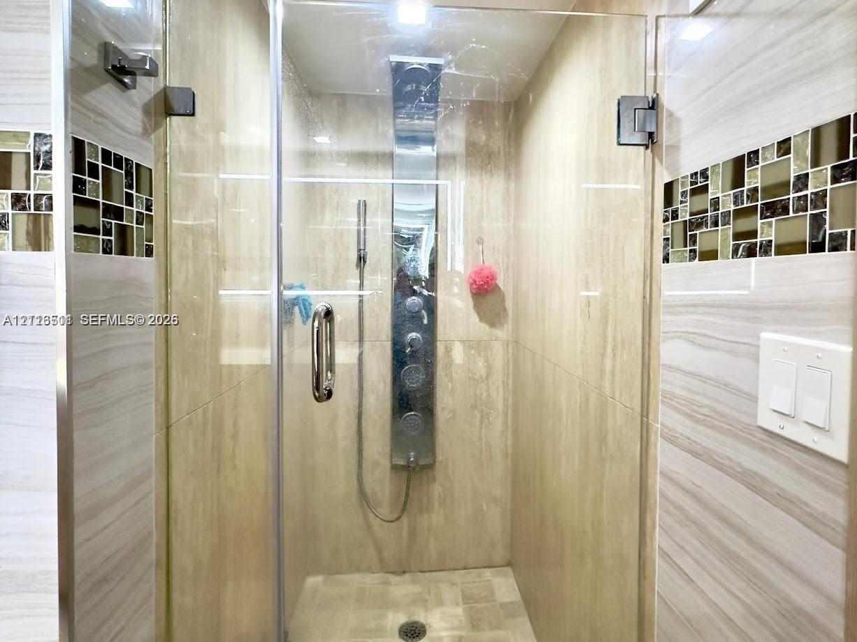 Walk-in shower featuring a frameless glass door, wall-mounted body spray panel, large-format tile surround, and decorative mosaic tile inlay