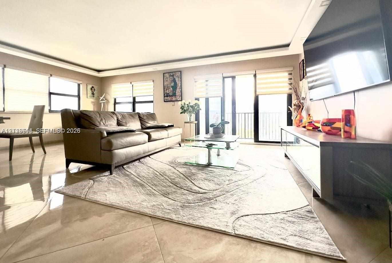 Living space featuring large-format tile flooring, recessed tray ceiling, multiple floor-to-ceiling windows with integrated roller shades, and sliding glass doors with balcony access