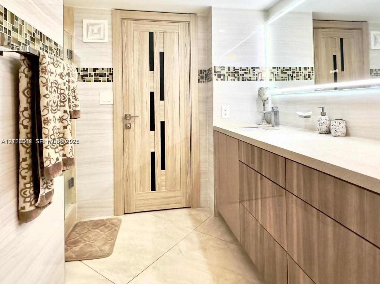 Bathroom featuring a floating vanity with wood-finish cabinetry, a large wall-to-wall mirror with integrated lighting, glass-tile decorative inlay, and a modern interior door with vertical dark-finish glass accents