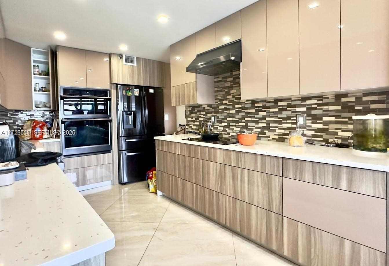 Kitchen featuring high-gloss cabinetry with wood-finish lower panels, solid surface countertops, a mosaic tile backsplash, and built-in double ovens