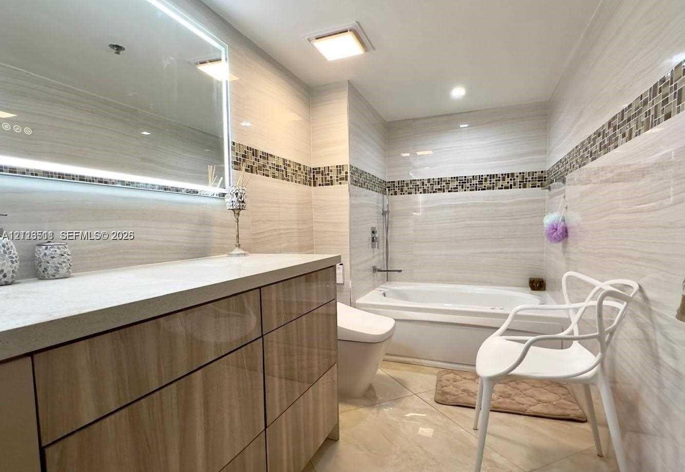 Bathroom featuring a gloss-finish wood cabinetry vanity, quartz countertop, illuminated wall mirror, integrated tile mosaic accents, and tile flooring