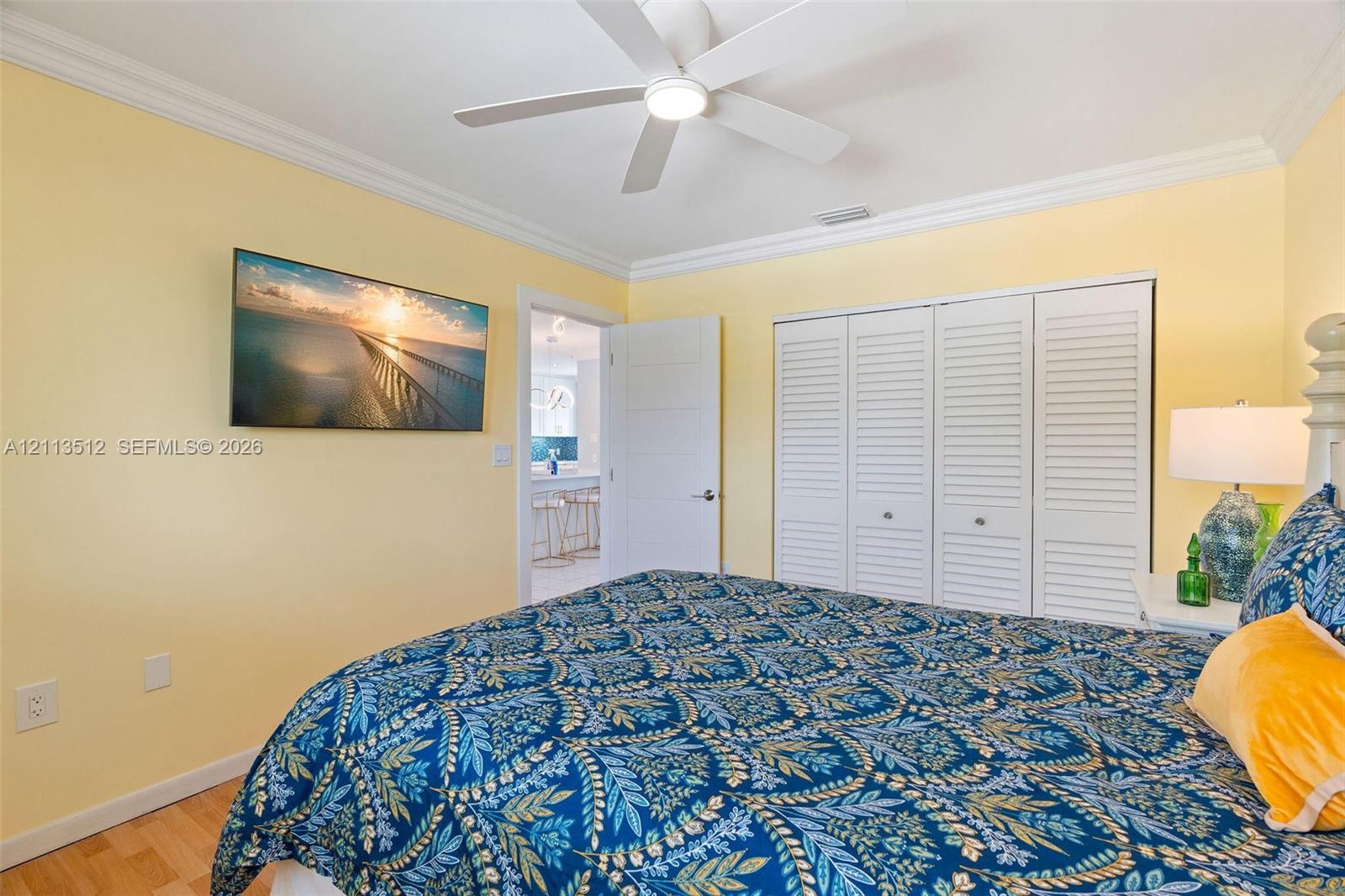 Room featuring white crown molding, a ceiling-mounted fan with integrated lighting, wood-finish flooring, and a triple-panel louvered closet door