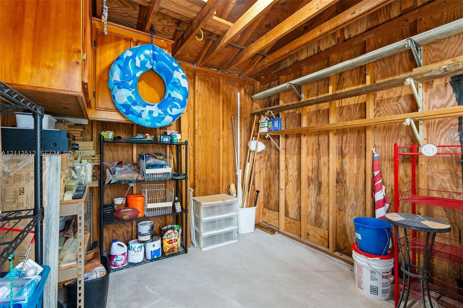 Storage area featuring wood-paneled walls, exposed wood ceiling rafters, wall-mounted shelving, and built-in upper cabinetry