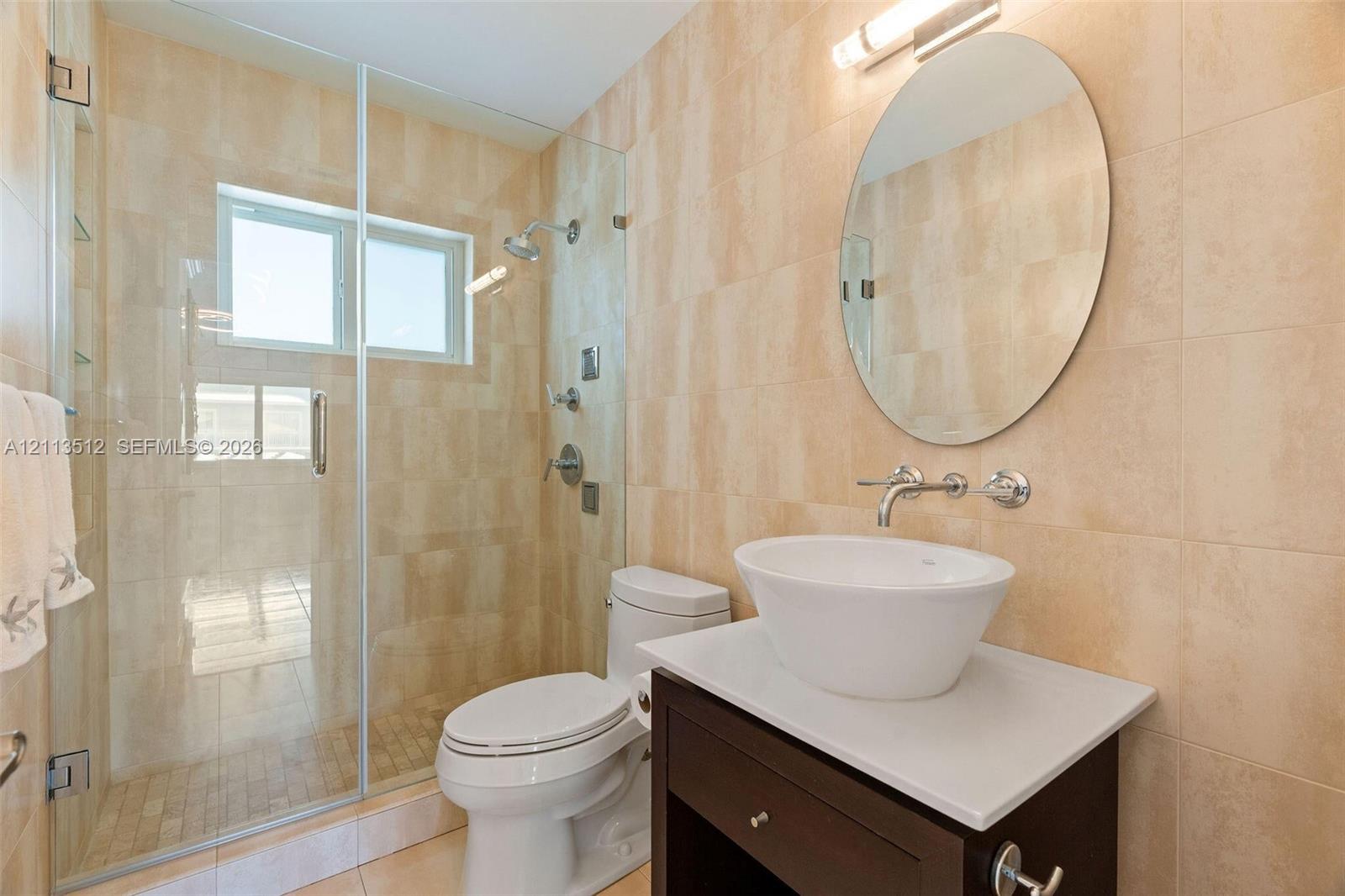 Bathroom featuring a vessel sink with a wall-mounted faucet, vanity with a solid-surface countertop, walk-in shower with a glass enclosure, and wall-to-wall ceramic tile finishes