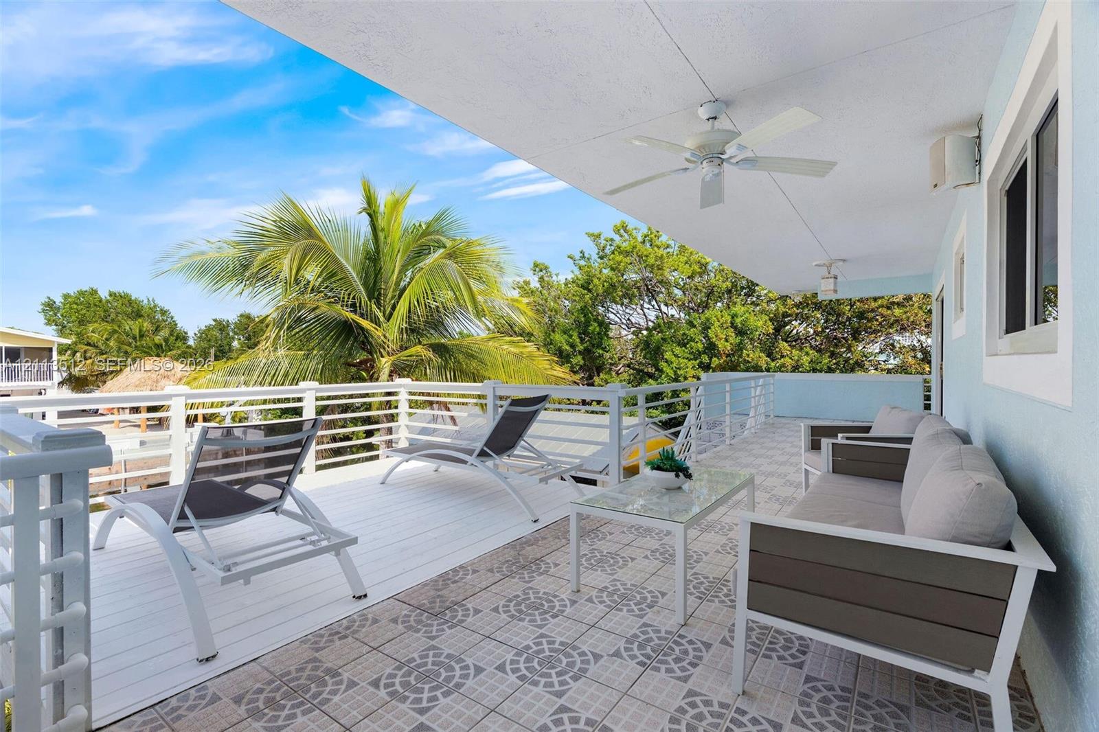 Covered balcony featuring white deck flooring, geometric patterned tile, white metal railings, and a ceiling-mounted fan