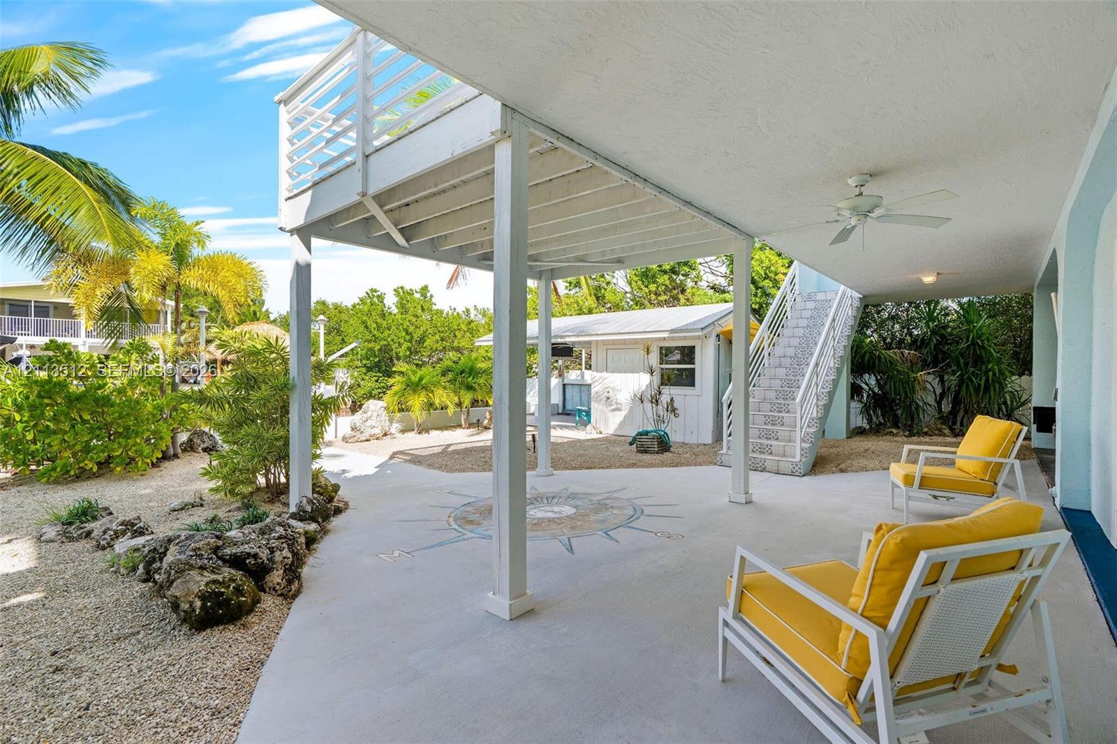 Covered ground-level patio featuring a decorative nautical compass inlay, elevated upper balcony, integrated ceiling fan, and staircase with metal railings