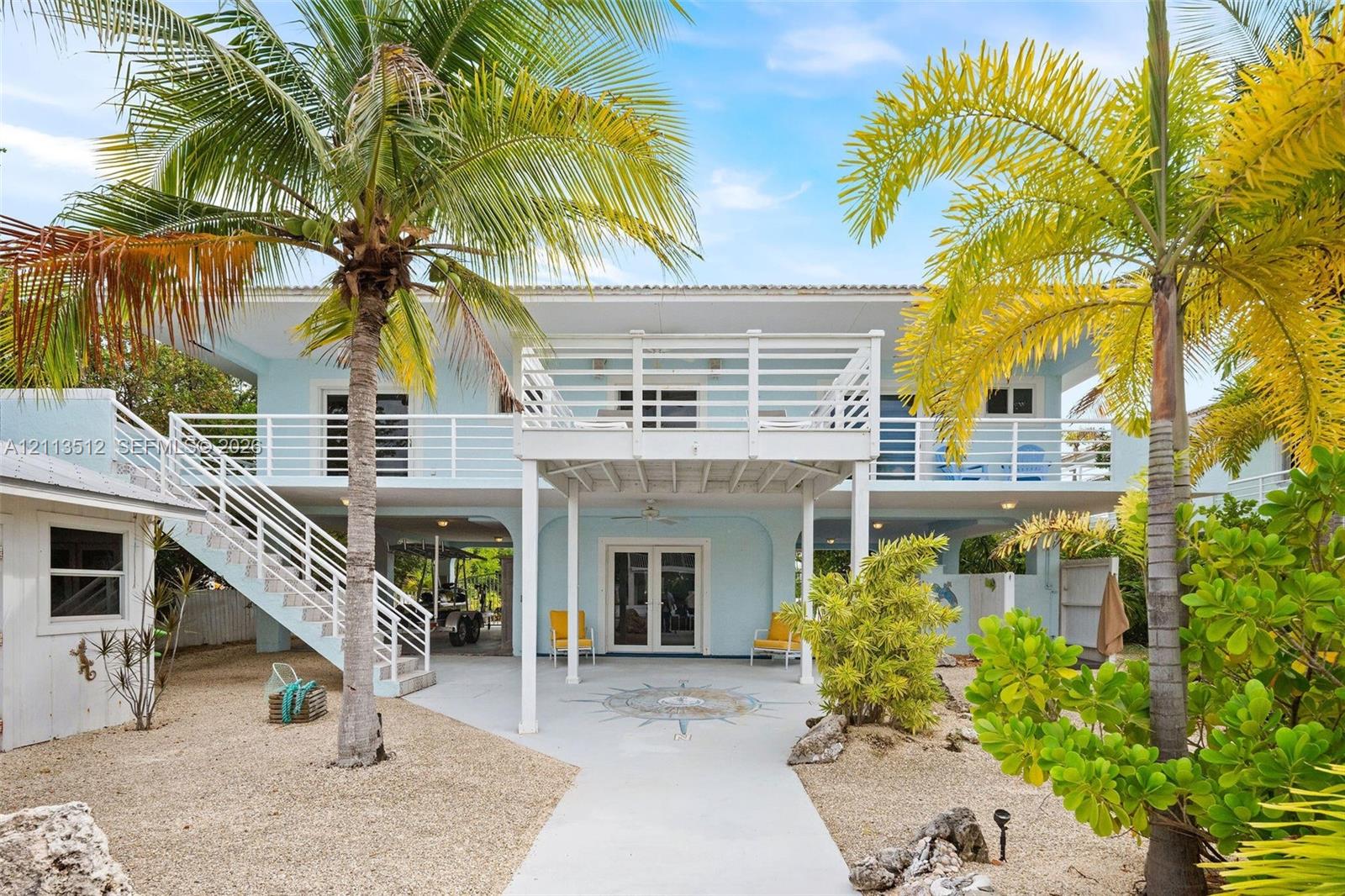 Elevated two-story residence featuring a ground-level covered patio with a decorative stone compass medallion, an expansive second-story balcony with white horizontal railings, an external staircase, and mature palm landscaping