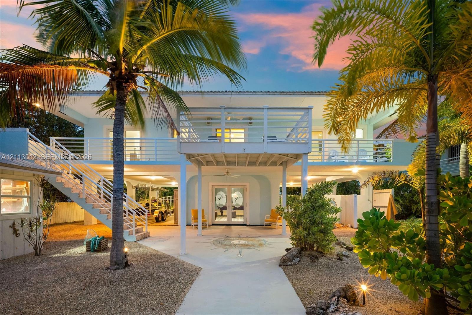 Two-story coastal residence featuring a stilt-construction design, a full-length elevated balcony with horizontal railings, an exterior staircase, and a ground-level covered patio with decorative stonework
