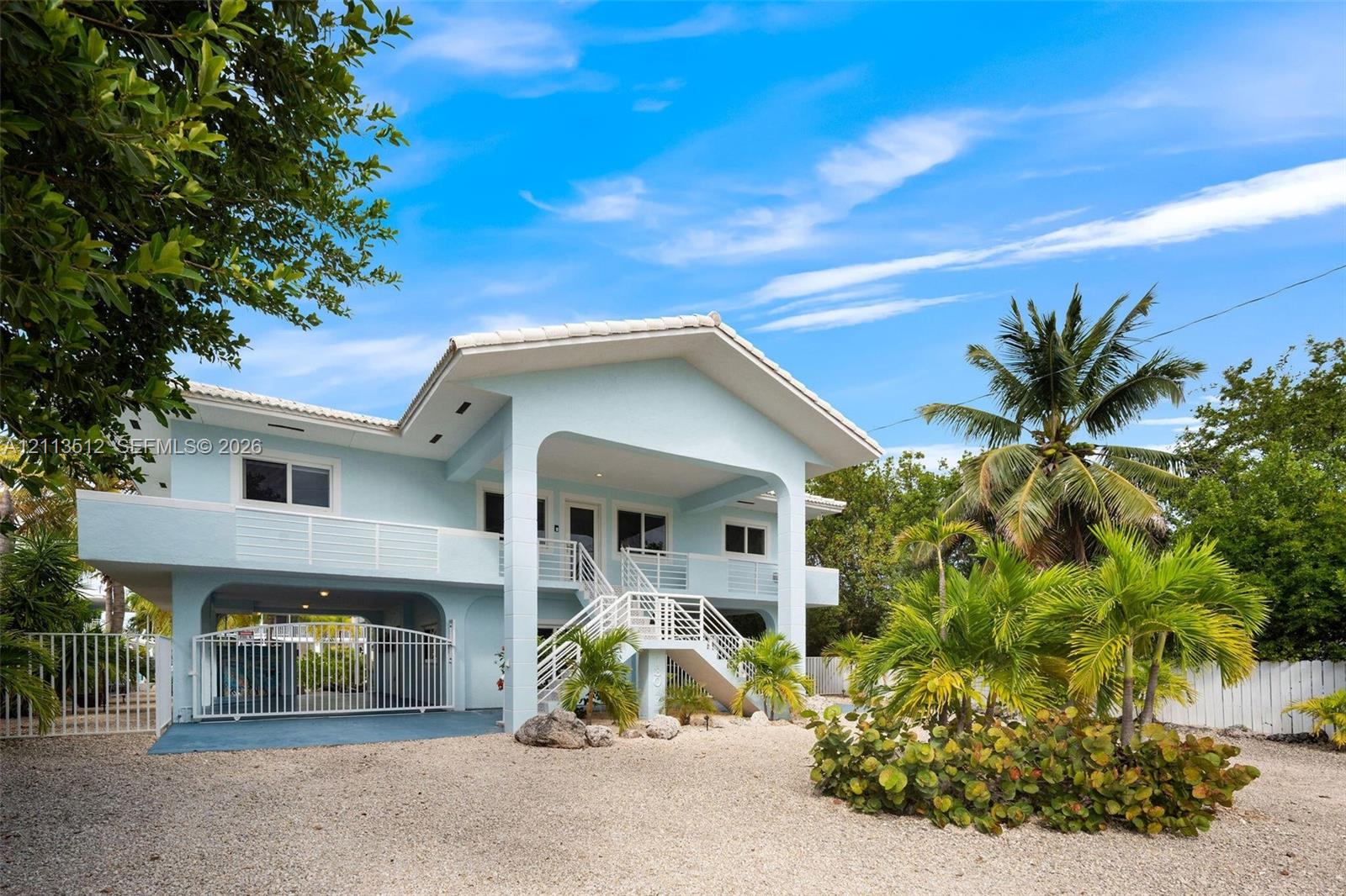 Elevated residence featuring a covered front porch, gated ground-level parking, metal railings, and a gravel driveway with mature landscaping