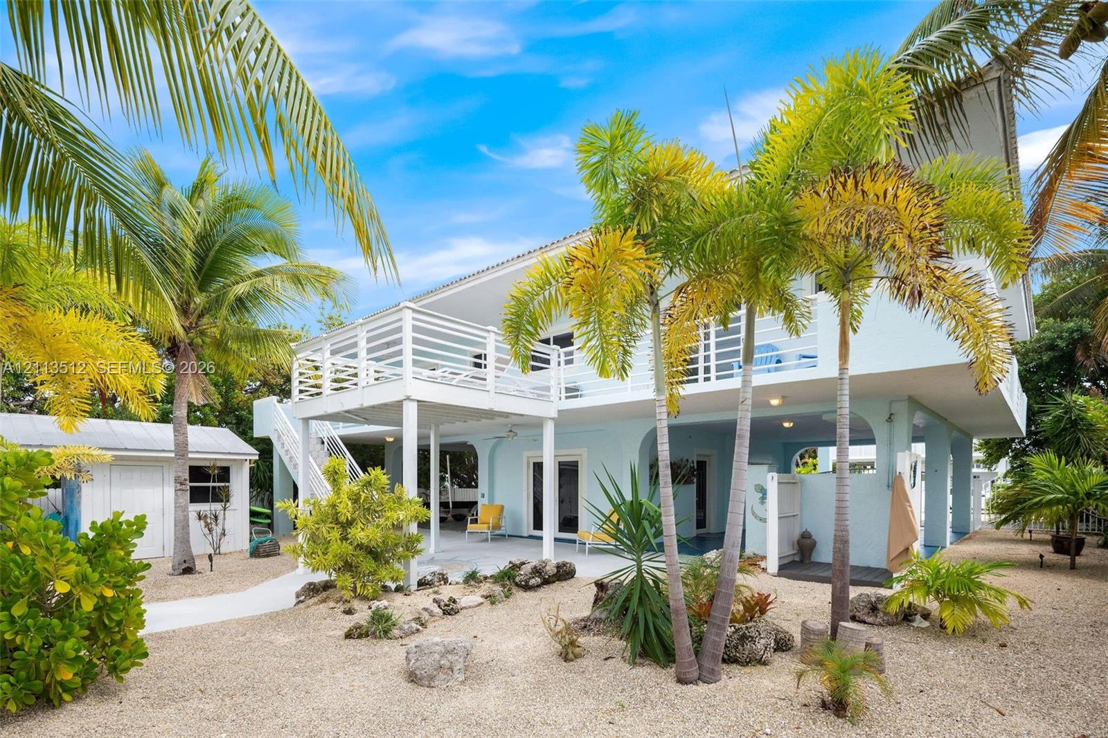 Elevated home exterior featuring a raised wooden balcony, covered ground-level patio, light-toned siding, and surrounding xeriscaped grounds with mature palm trees