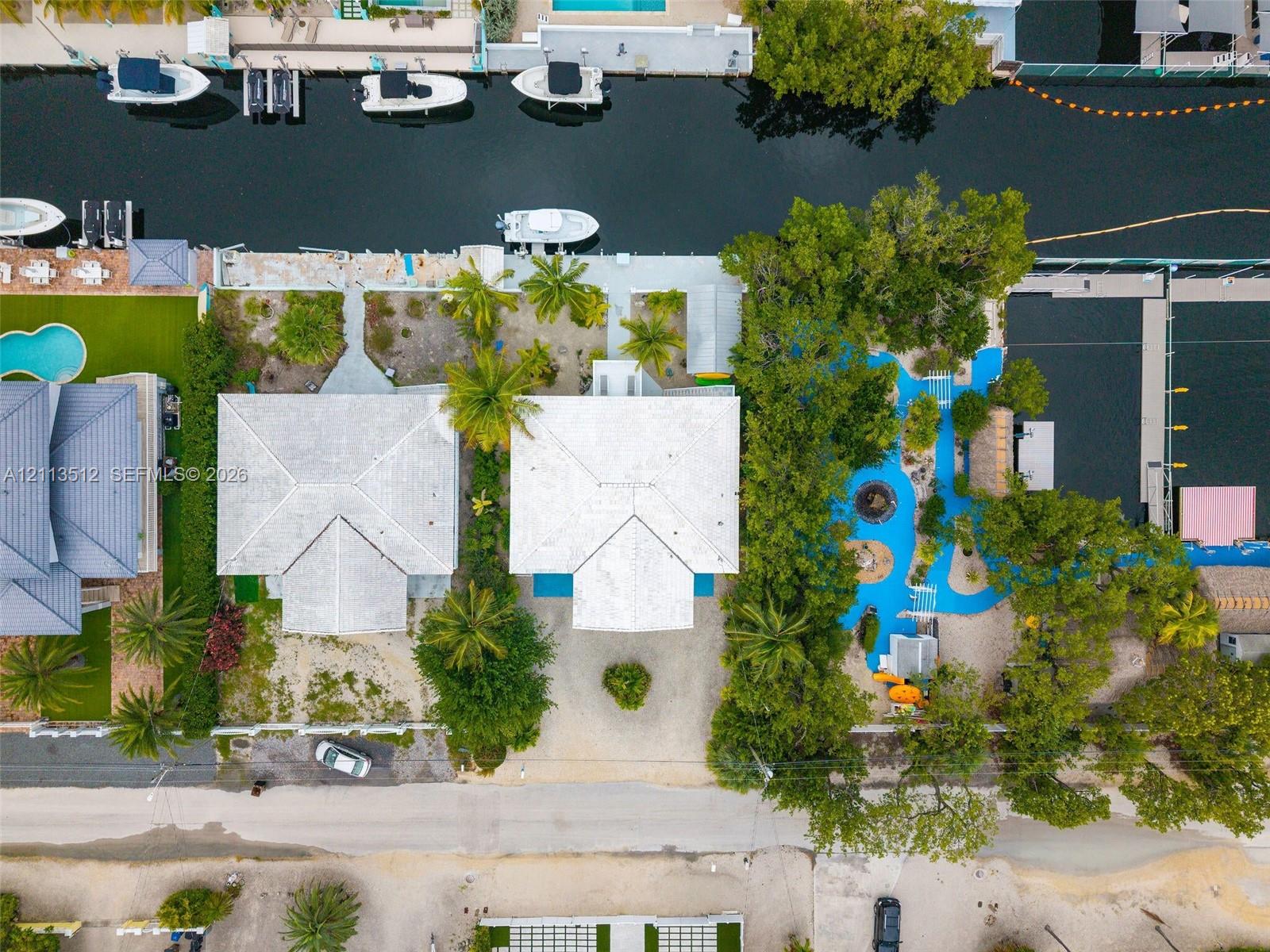 Aerial view of a waterfront property featuring direct canal access, a private boat slip, paved driveway, mature tropical landscaping, and a blue-surfaced outdoor recreation area
