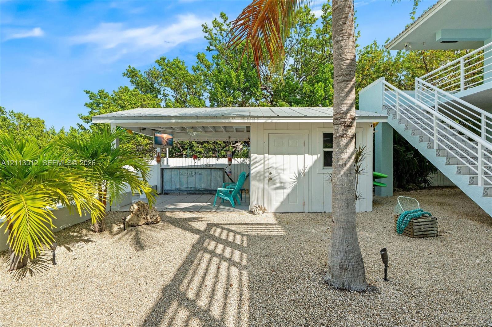 Outdoor covered bar area featuring a metal roof and concrete structure, surrounded by gravel grounds with mature palm trees and exterior stairs with white railing