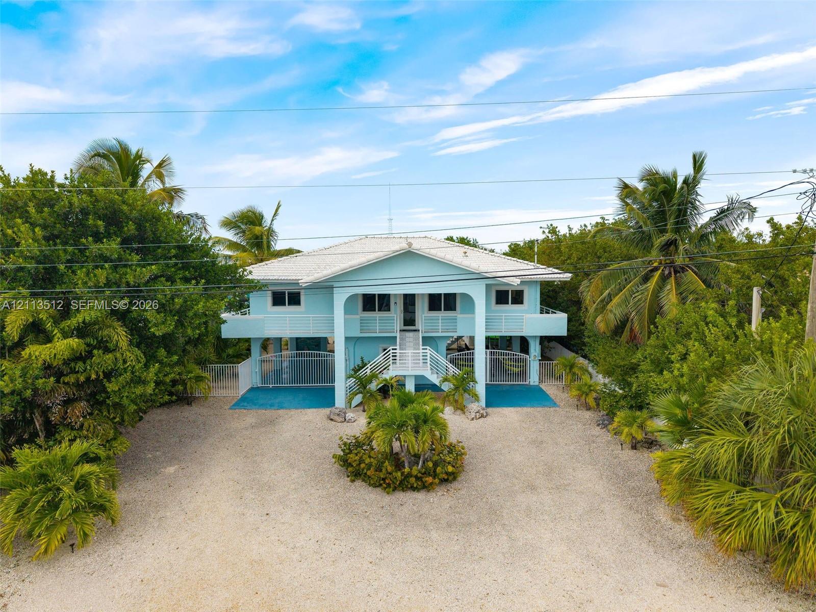 Elevated coastal residence featuring a metal roof, wraparound balcony, central staircase with white railing, and gravel driveway