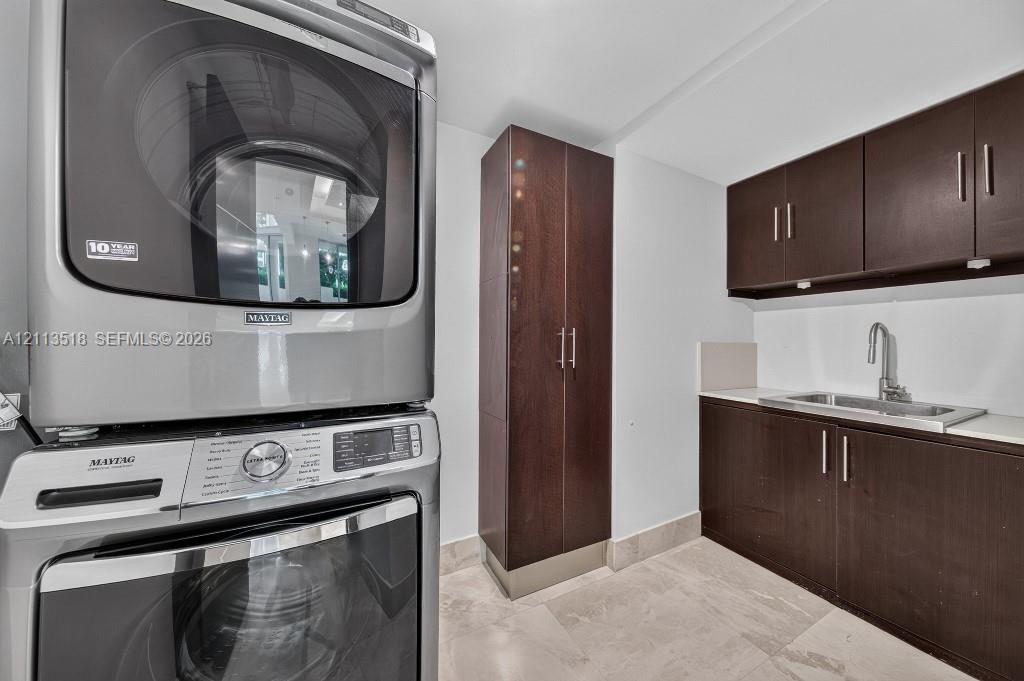 Laundry area featuring dark wood-finish cabinetry, a tall pantry unit, a stainless steel undermount sink with a gooseneck faucet, light-toned stone-finish flooring, and a stacked front-loading washer and dryer set