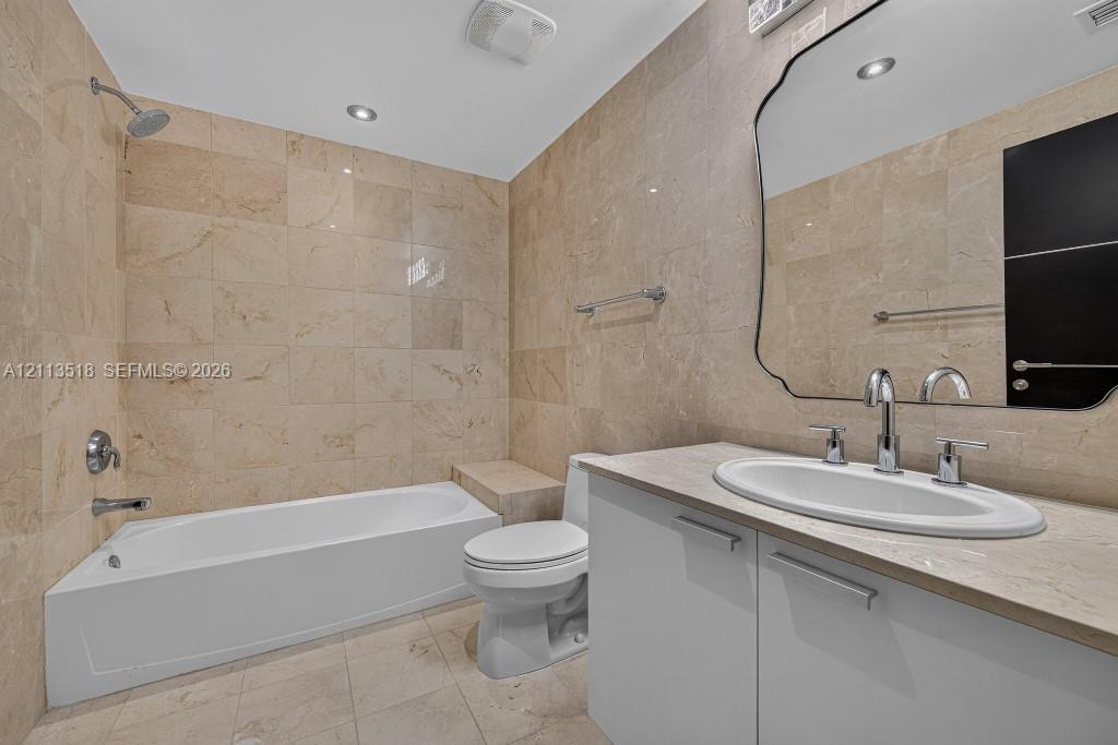 Full-height natural stone wall tiling, white vanity with an integrated countertop, oval undermount sink, decorative wall mirror, and built-in tub surround