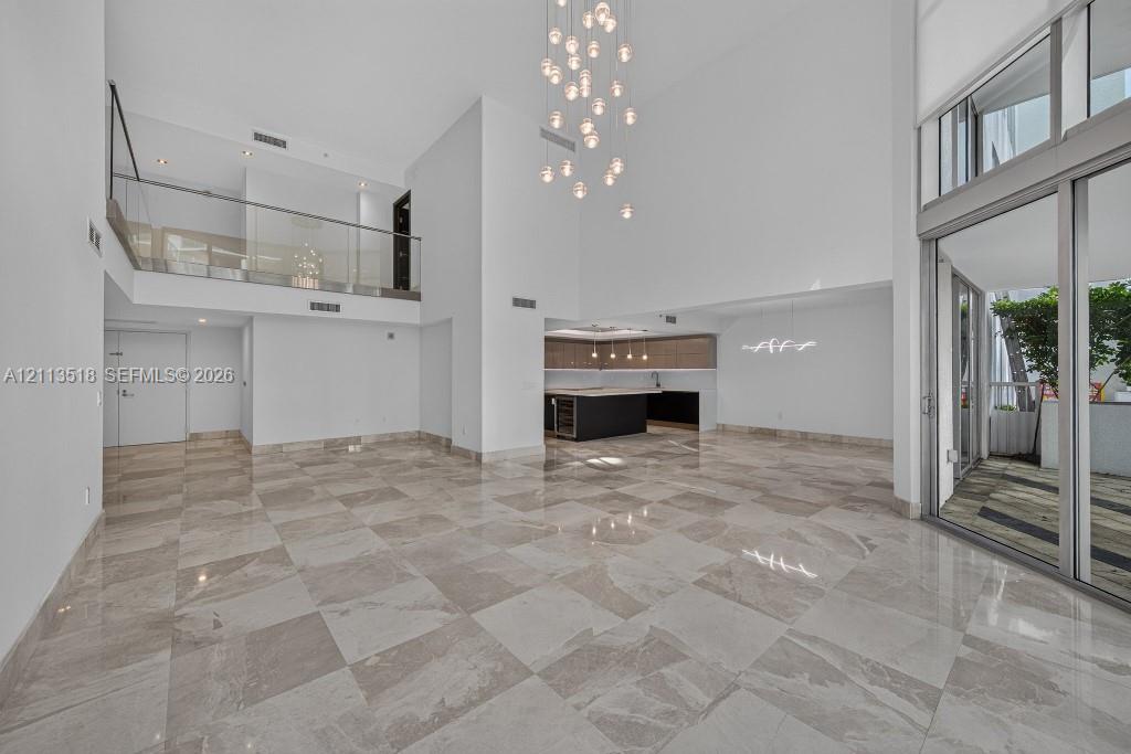 Open-concept interior featuring double-height ceilings, stone-finish tile flooring, a glass-panel mezzanine railing, multi-light pendant chandelier, and expansive floor-to-ceiling sliding glass doors Open-concept interior featuring double-height ceilings, stone-finish tile flooring, a glass-panel mezzanine railing, multi-light pendant chandelier, and expansive floor-to-ceiling sliding glass doors