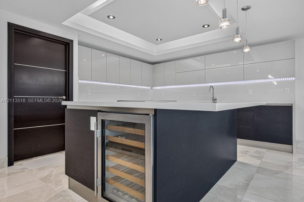 Kitchen featuring high-gloss cabinetry, a central island with a built-in wine refrigerator, stone countertops, recessed tray ceiling lighting, and tile flooring