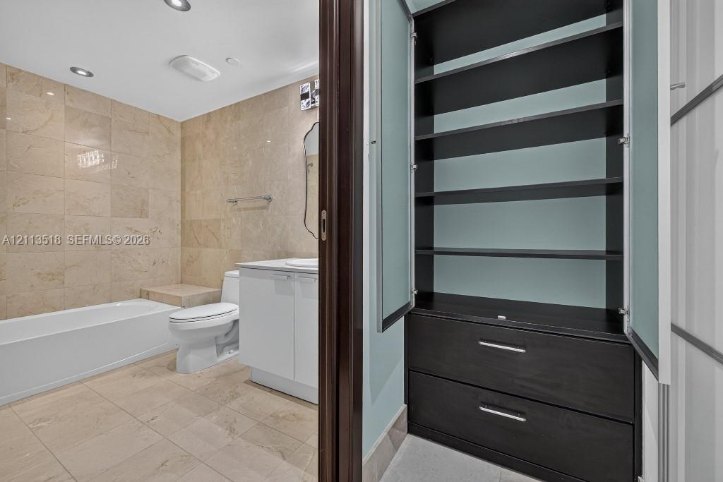 Built-in cabinetry featuring dark wood-finish shelving and drawers with frosted glass doors, complemented by a bathroom finished with stone-tile wall and floor surfacing, a white vanity cabinet, and a built-in bathtub