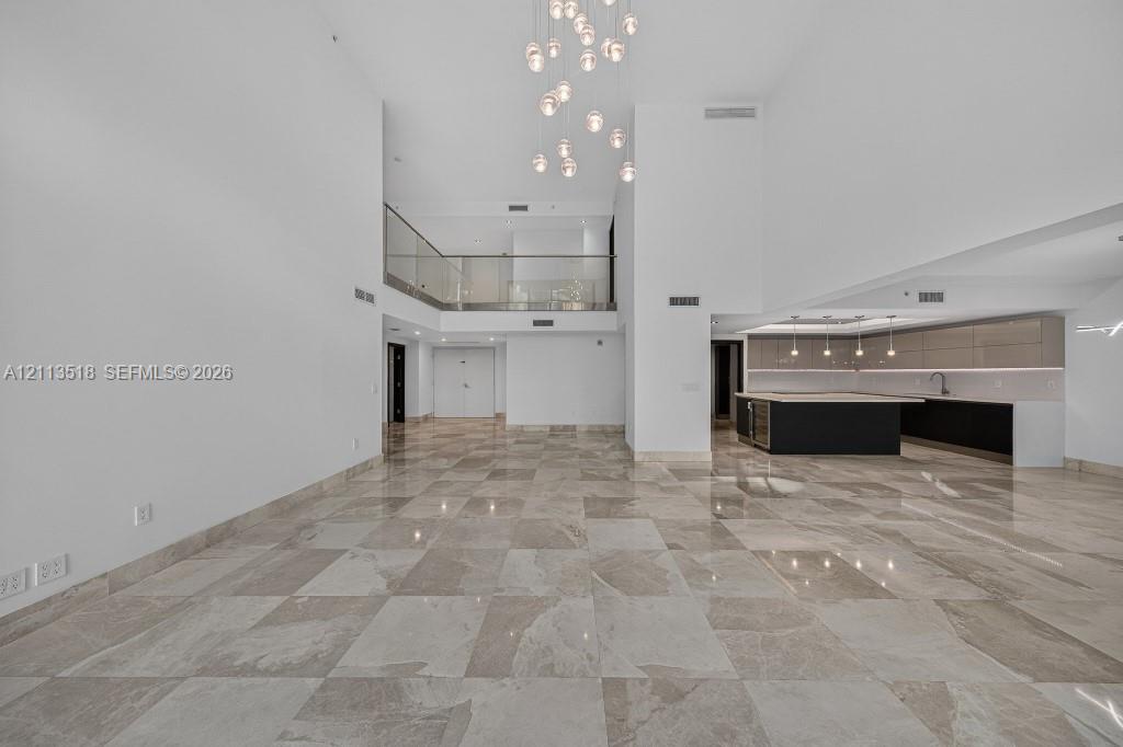 Expansive interior featuring high ceilings, stone flooring, a multi-tiered glass pendant chandelier, a glass-railed mezzanine, and kitchen cabinetry with integrated lighting Expansive interior featuring high ceilings, stone flooring, a multi-tiered glass pendant chandelier, a glass-railed mezzanine, and kitchen cabinetry with integrated lighting