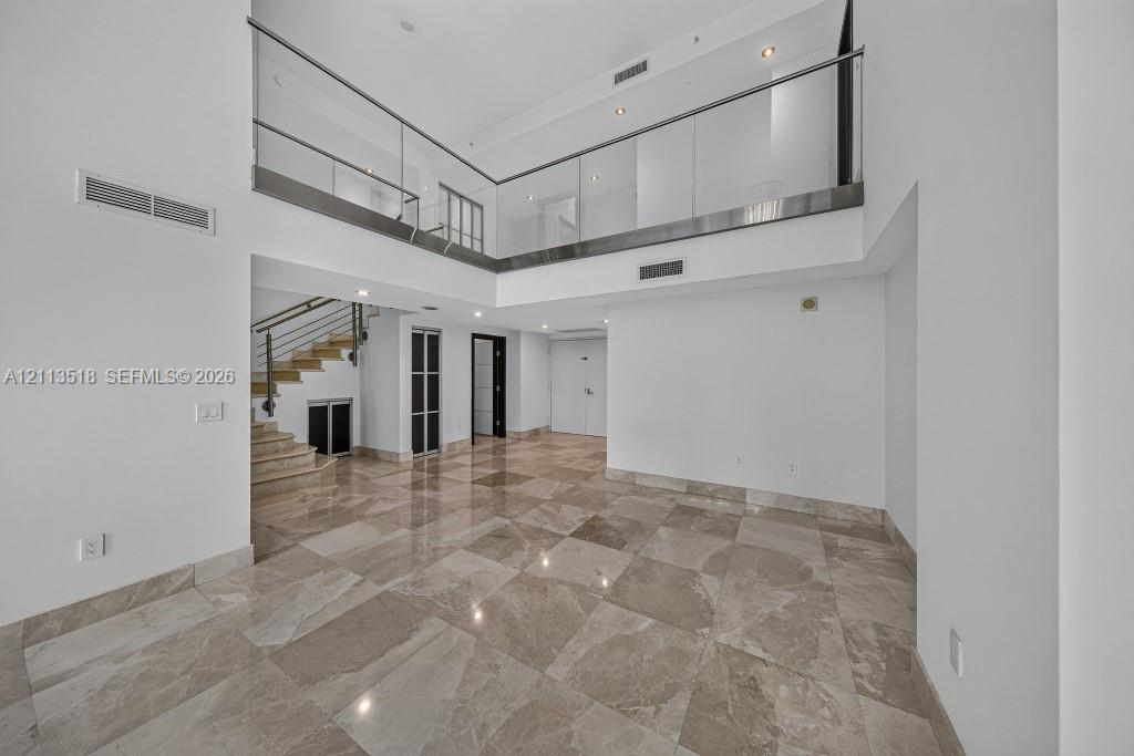 Double-height ceiling, polished stone flooring, glass panel mezzanine railing, recessed lighting, and staircase with metal handrail