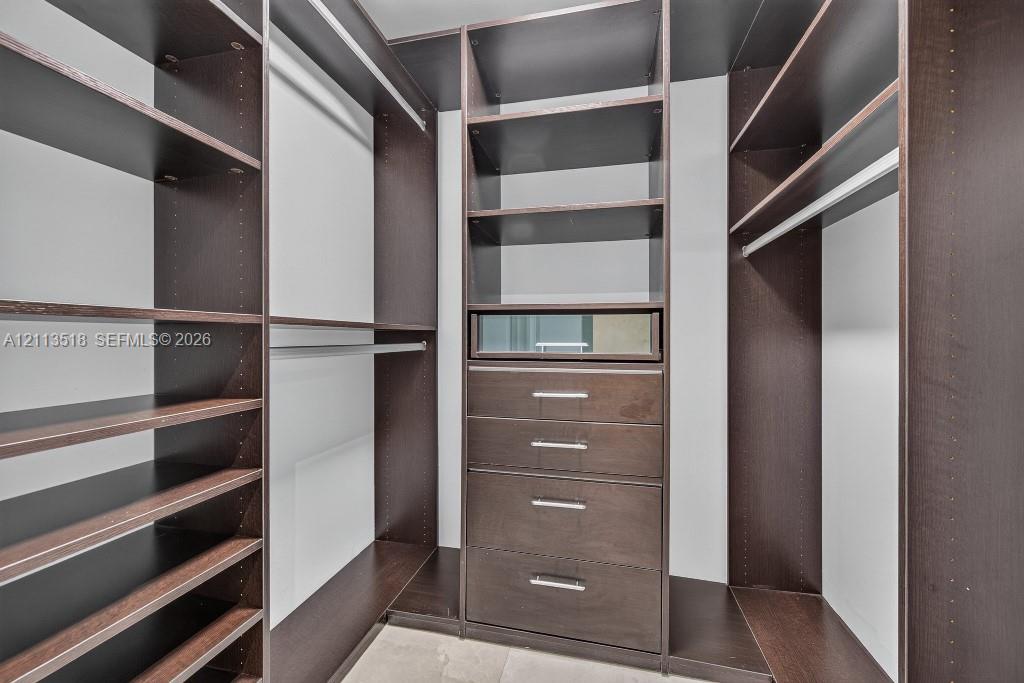 Custom walk-in storage system featuring dark wood-finish cabinetry, built-in shelving, integrated hanging rods, and a centralized unit with four drawers and a glass-front display compartment