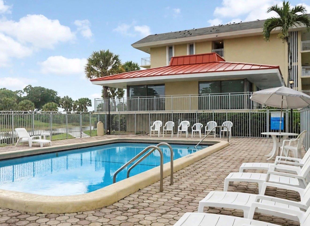Swimming pool with concrete paver decking, metal perimeter fencing, and a structure featuring a red metal roof Swimming pool with concrete paver decking, metal perimeter fencing, and a structure featuring a red metal roof