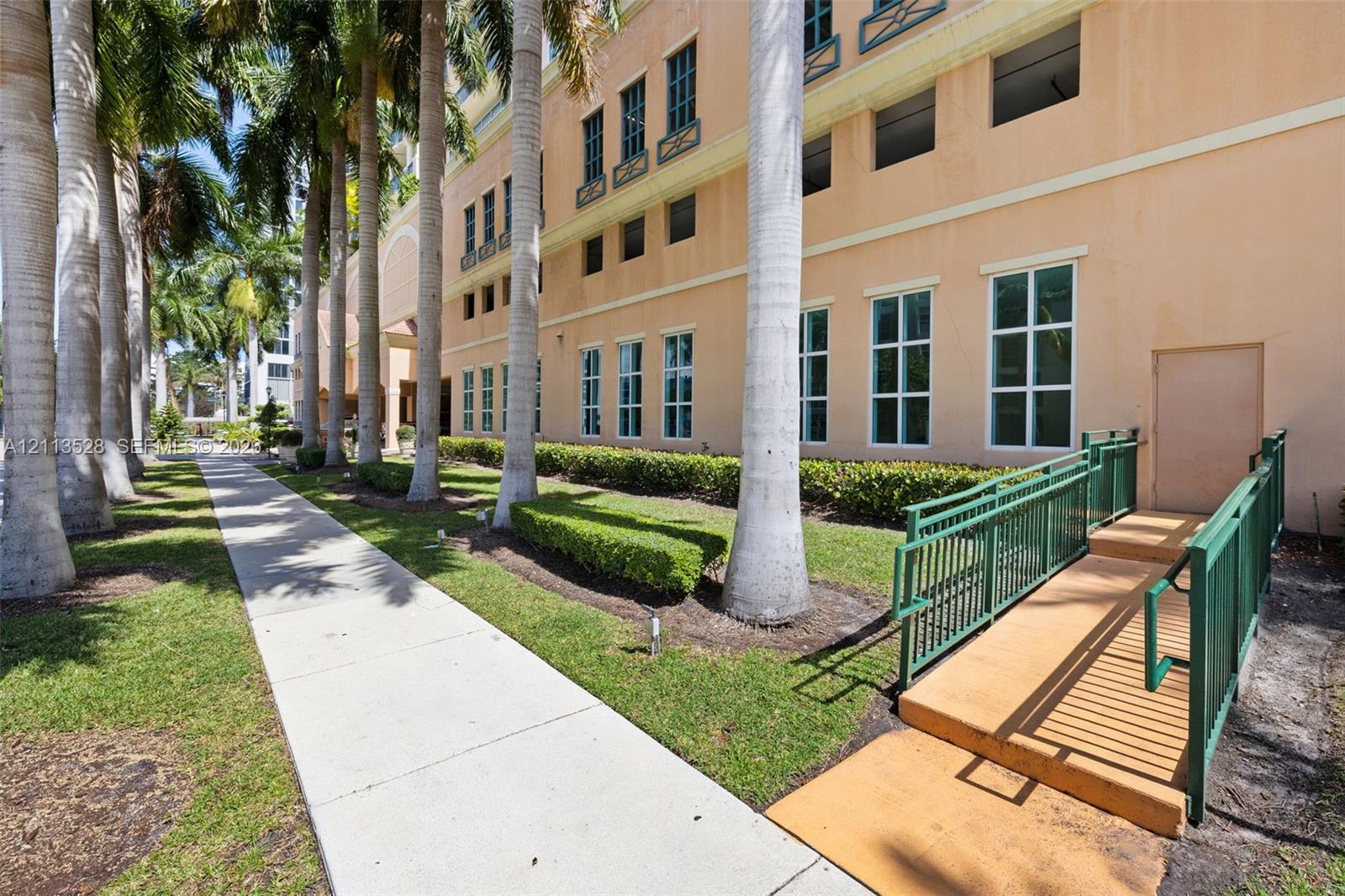 Exterior facade featuring multi-story stucco construction, manicured landscaping with mature palm trees, multi-pane windows, and a dedicated ramp with metal handrails