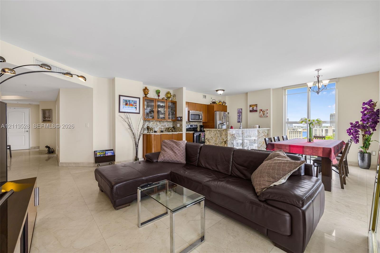 Open-concept main living area featuring light-toned tile flooring, built-in wooden cabinetry with glass-front uppers, a kitchen island with stone countertop, and expansive floor-to-ceiling windows