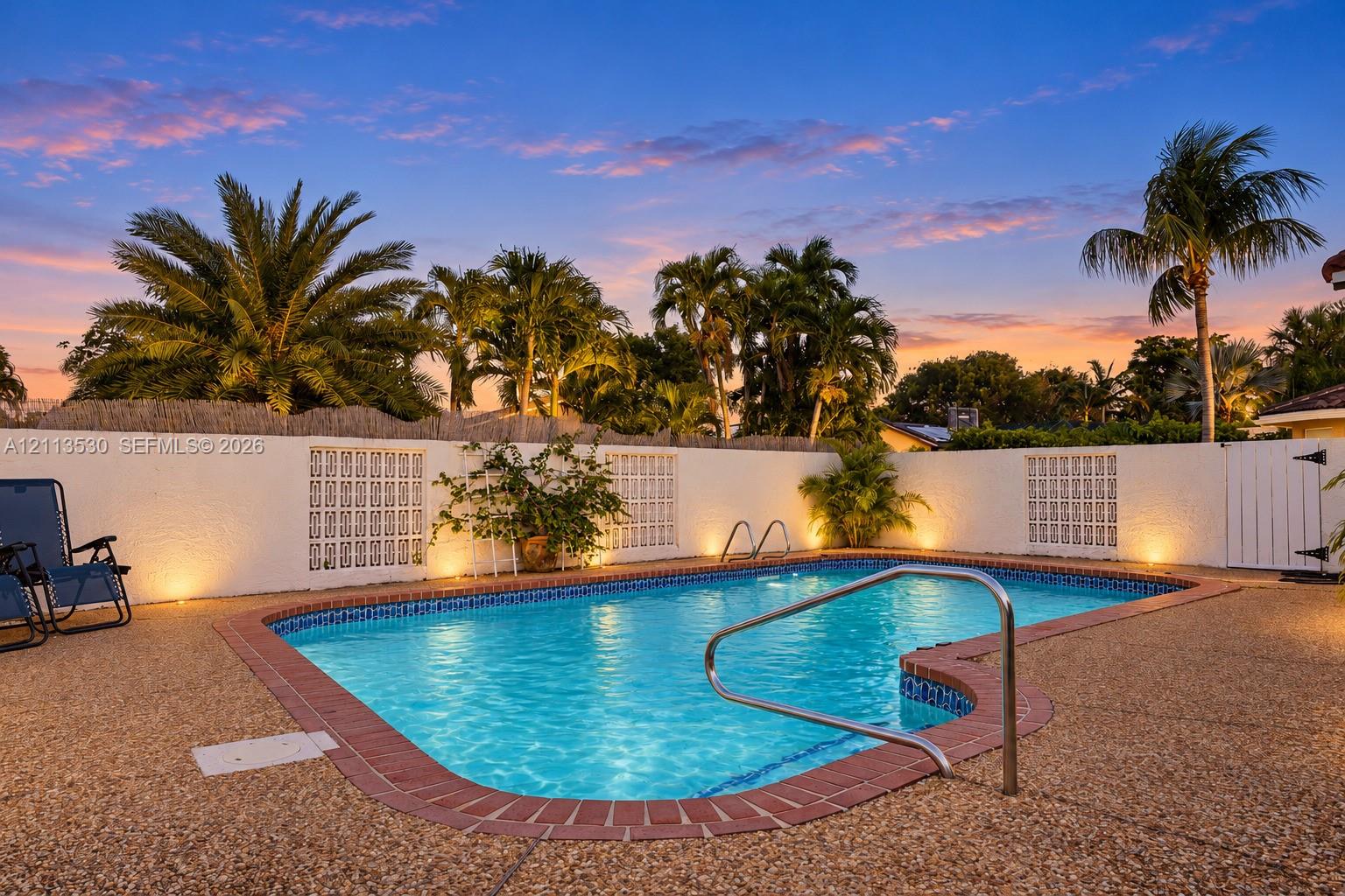 In-ground swimming pool featuring a brick-tiled coping edge, a metal handrail, and a textured stone-finish patio surround enclosed by a masonry privacy wall with decorative lattice panels
