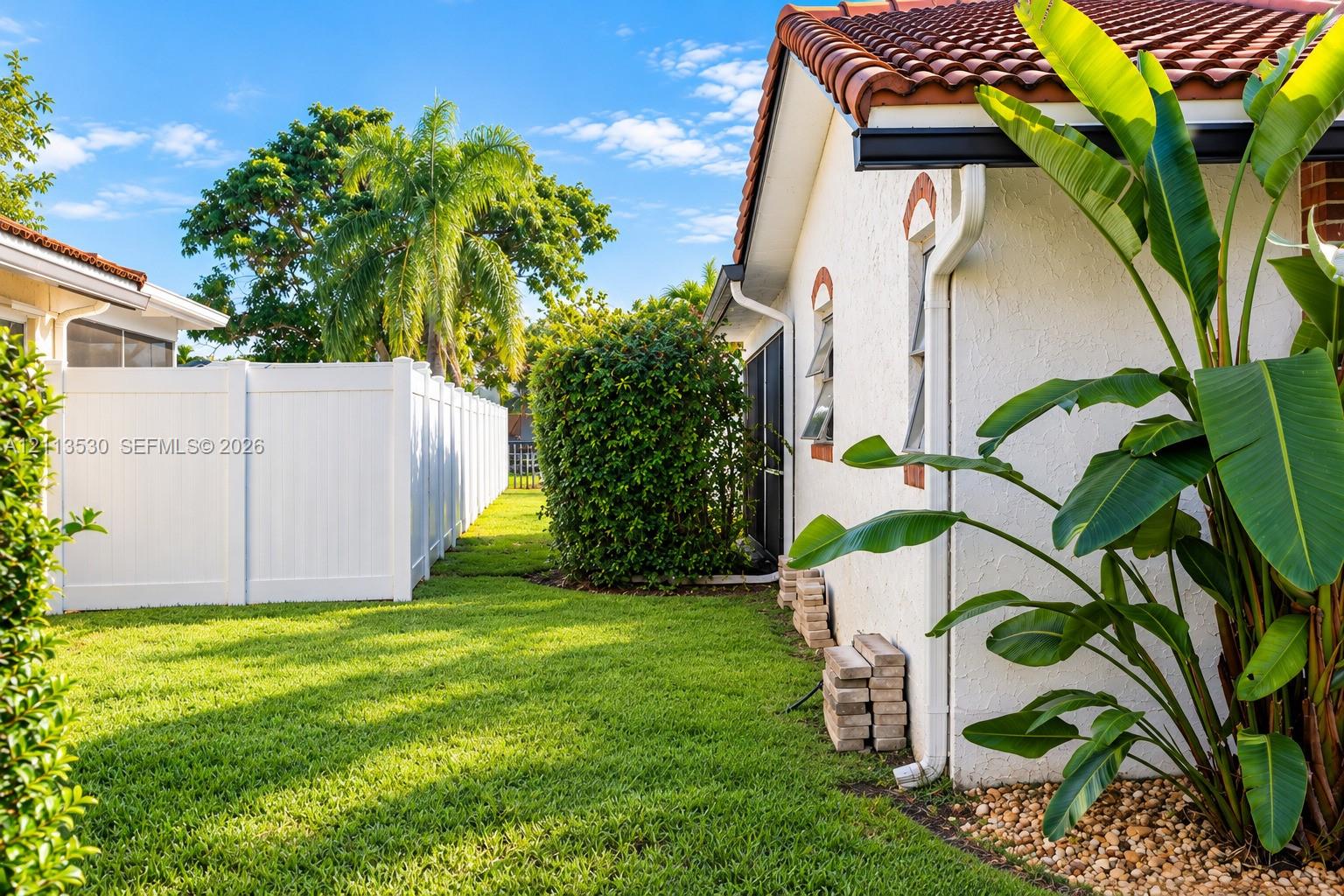 Stucco exterior with terracotta-style clay tile roofing, white vinyl privacy fencing, and landscaped side yard