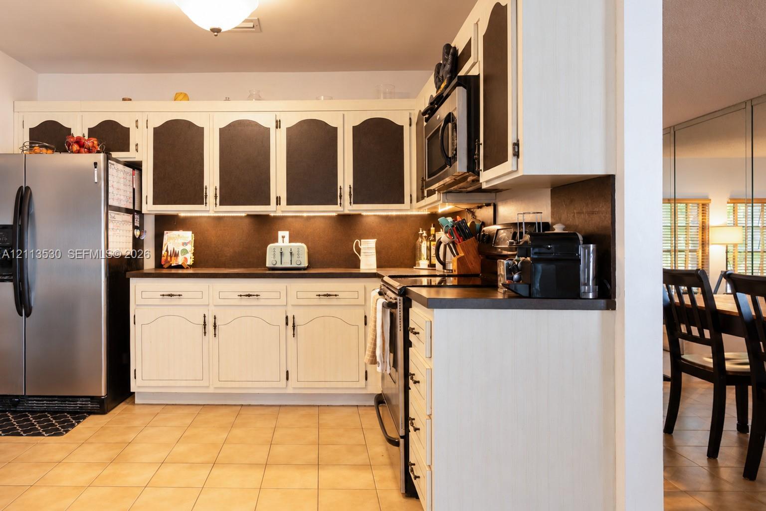 Kitchen featuring custom two-tone cabinetry, solid surface countertops, under-cabinet lighting, stainless steel appliances, and ceramic tile flooring