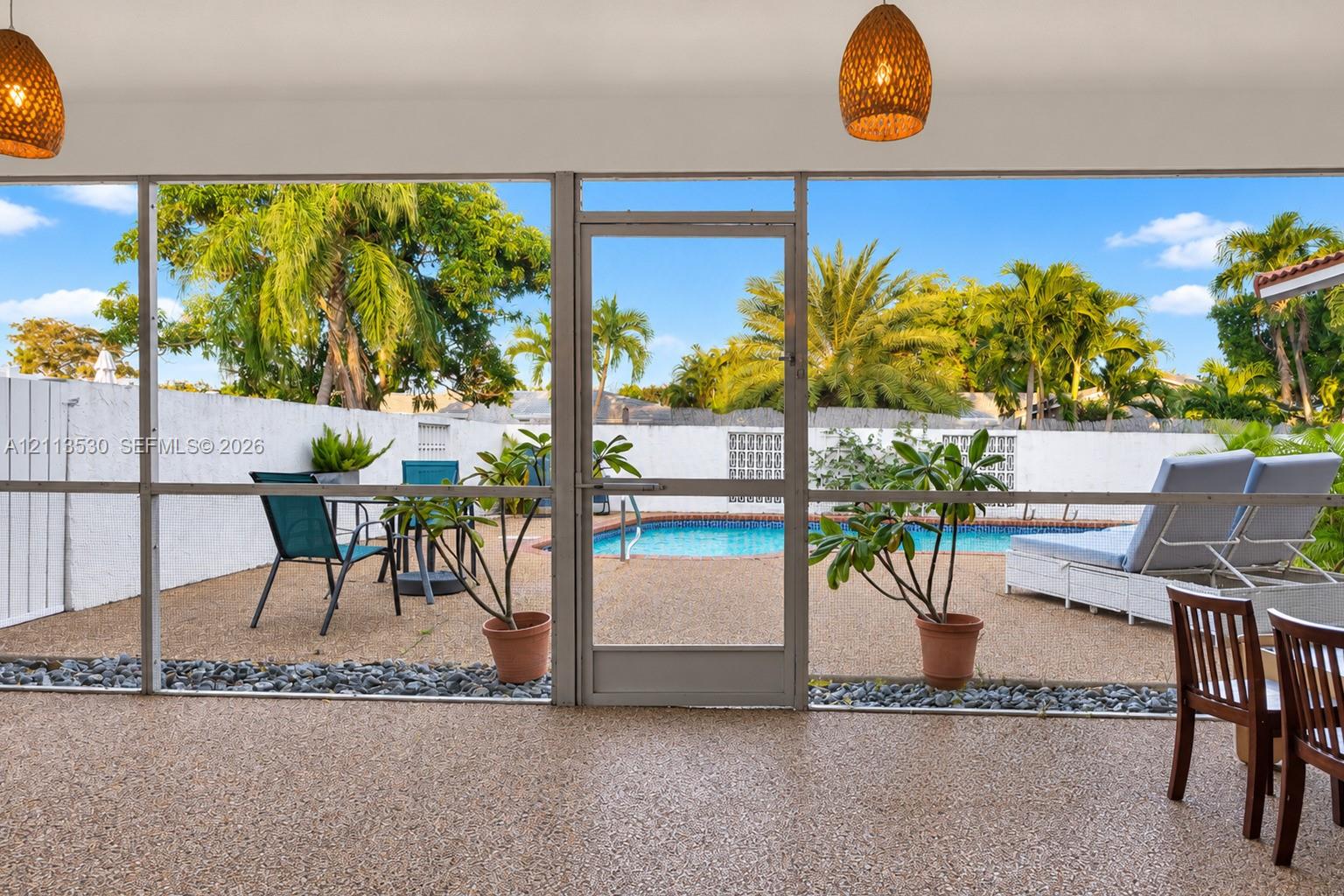 Screen-enclosed patio with textured aggregate flooring, dual woven-texture pendant light fixtures, and a direct entrance to an outdoor swimming pool area featuring a perimeter privacy wall