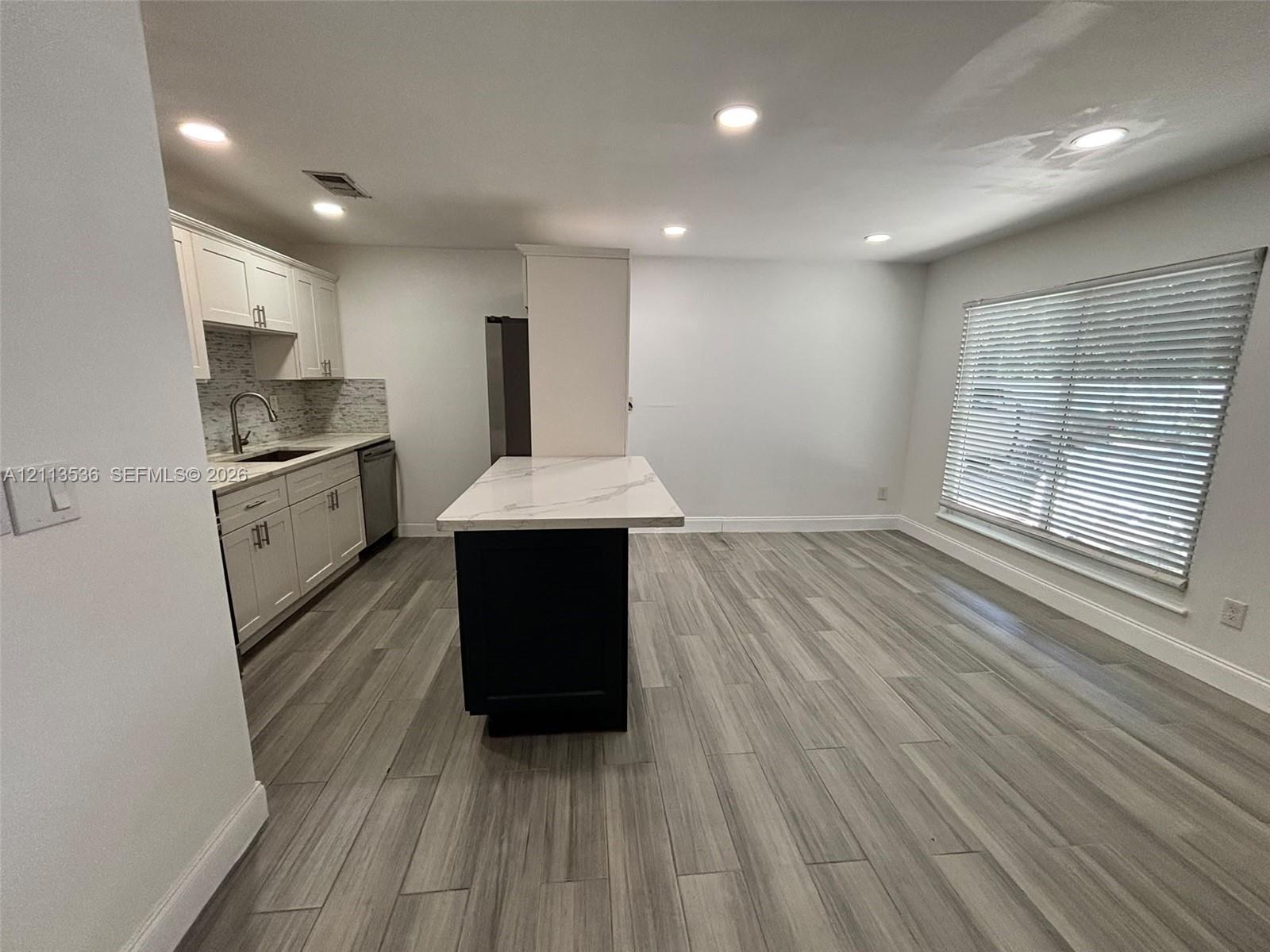 Kitchen featuring white cabinetry, a contrasting dark central island with a stone-finish countertop, wood-finish flooring, recessed lighting, and a window with blinds Kitchen featuring white cabinetry, a contrasting dark central island with a stone-finish countertop, wood-finish flooring, recessed lighting, and a window with blinds