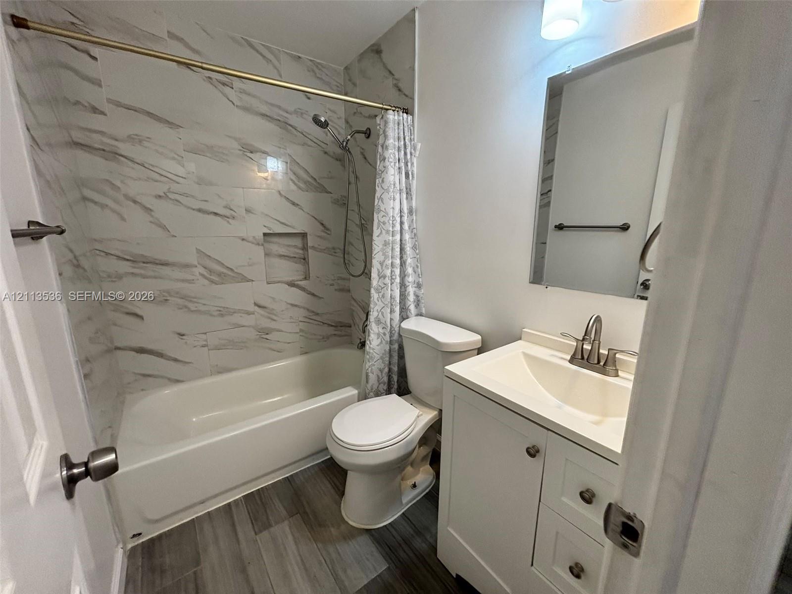 Bathroom featuring wood-finish flooring, white vanity with integrated basin, tub-shower combination with full-height stone-patterned tile surround, and built-in shower niche Bathroom featuring wood-finish flooring, white vanity with integrated basin, tub-shower combination with full-height stone-patterned tile surround, and built-in shower niche