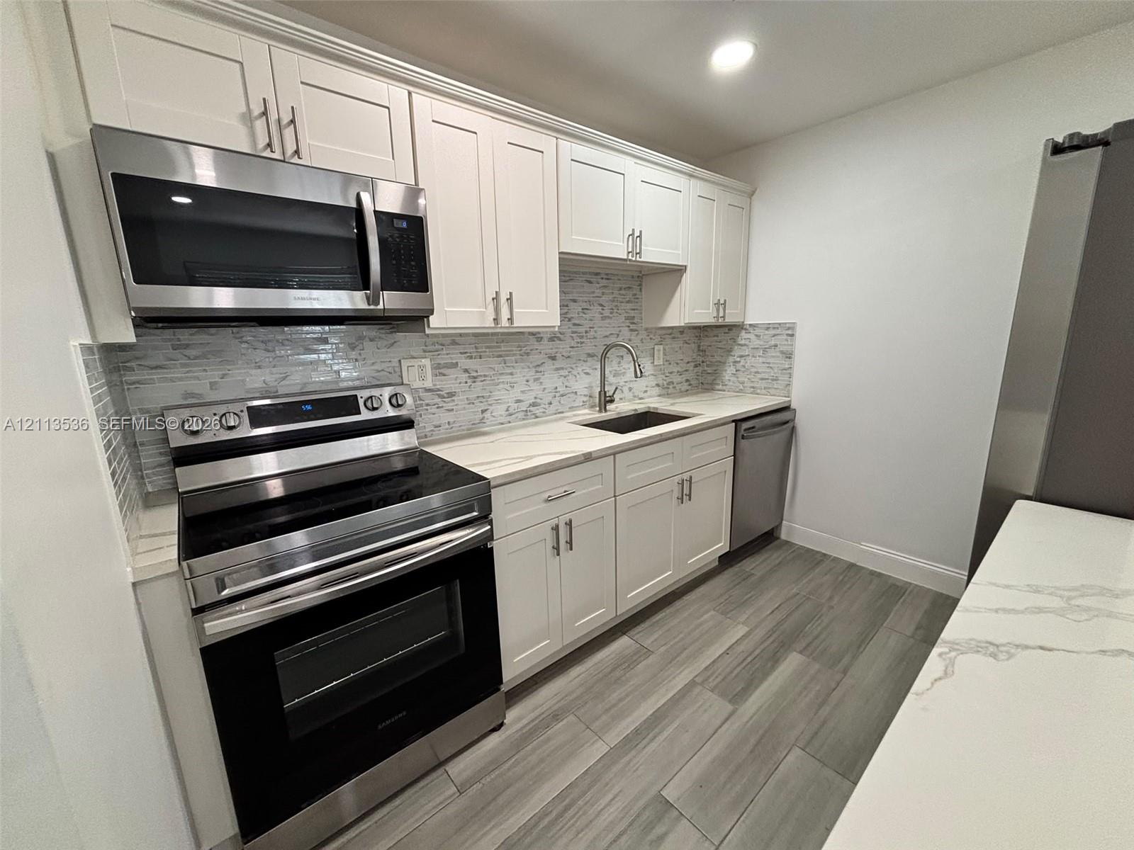 Kitchen featuring white Shaker-style cabinetry, stainless steel appliances, quartz countertops, a stone-tile backsplash, and wood-finish flooring Kitchen featuring white Shaker-style cabinetry, stainless steel appliances, quartz countertops, a stone-tile backsplash, and wood-finish flooring