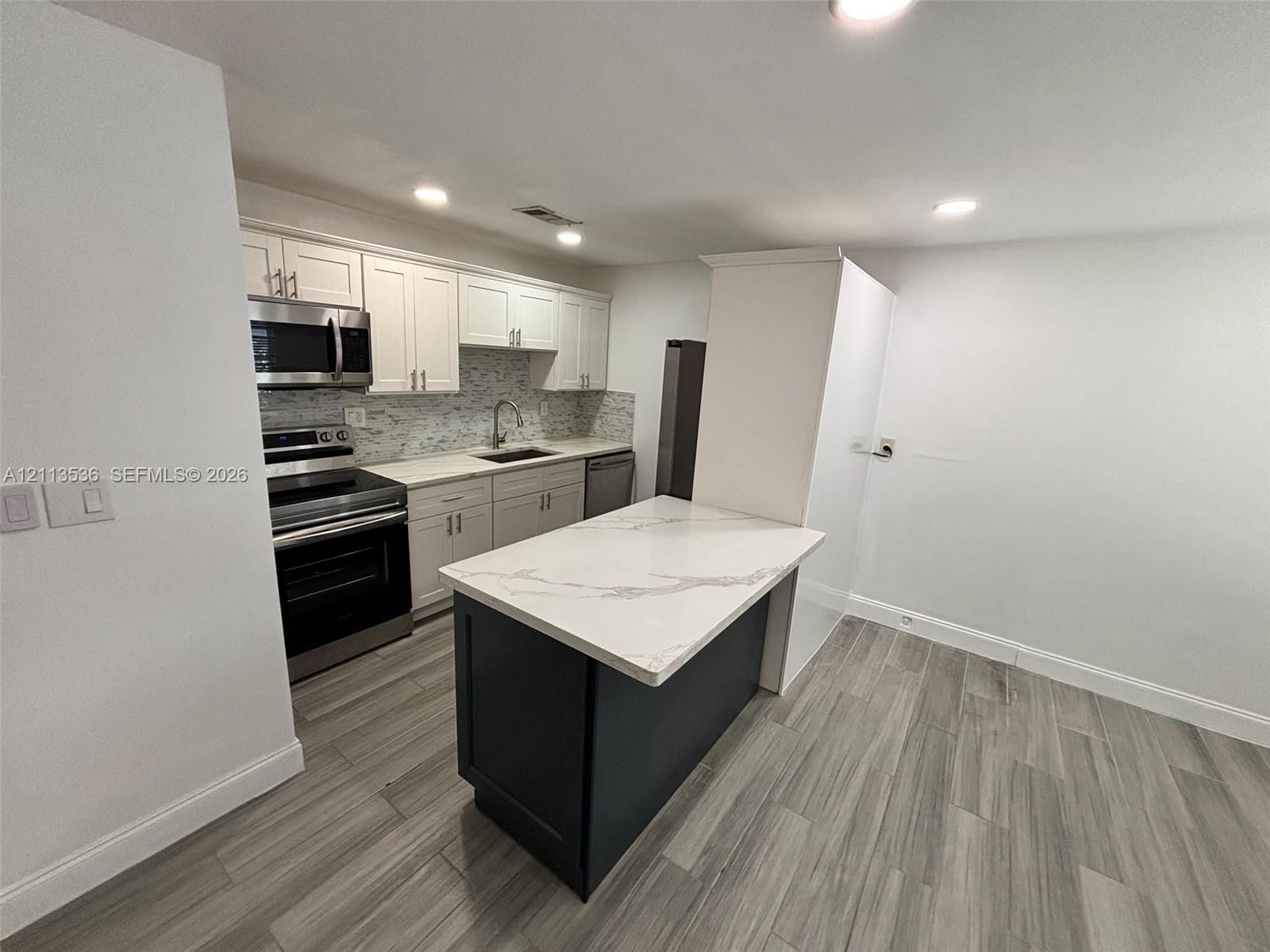 Kitchen featuring white cabinetry, an island with a stone-composite countertop, stainless steel appliances, a tile backsplash, and wood-finish flooring Kitchen featuring white cabinetry, an island with a stone-composite countertop, stainless steel appliances, a tile backsplash, and wood-finish flooring