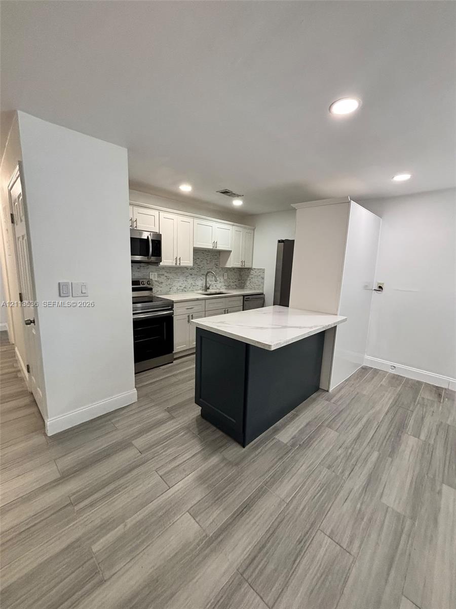 Kitchen featuring white cabinetry, stone-finish countertops, a contrasting dark-finish kitchen island, stainless steel appliances, and wood-finish flooring Kitchen featuring white cabinetry, stone-finish countertops, a contrasting dark-finish kitchen island, stainless steel appliances, and wood-finish flooring