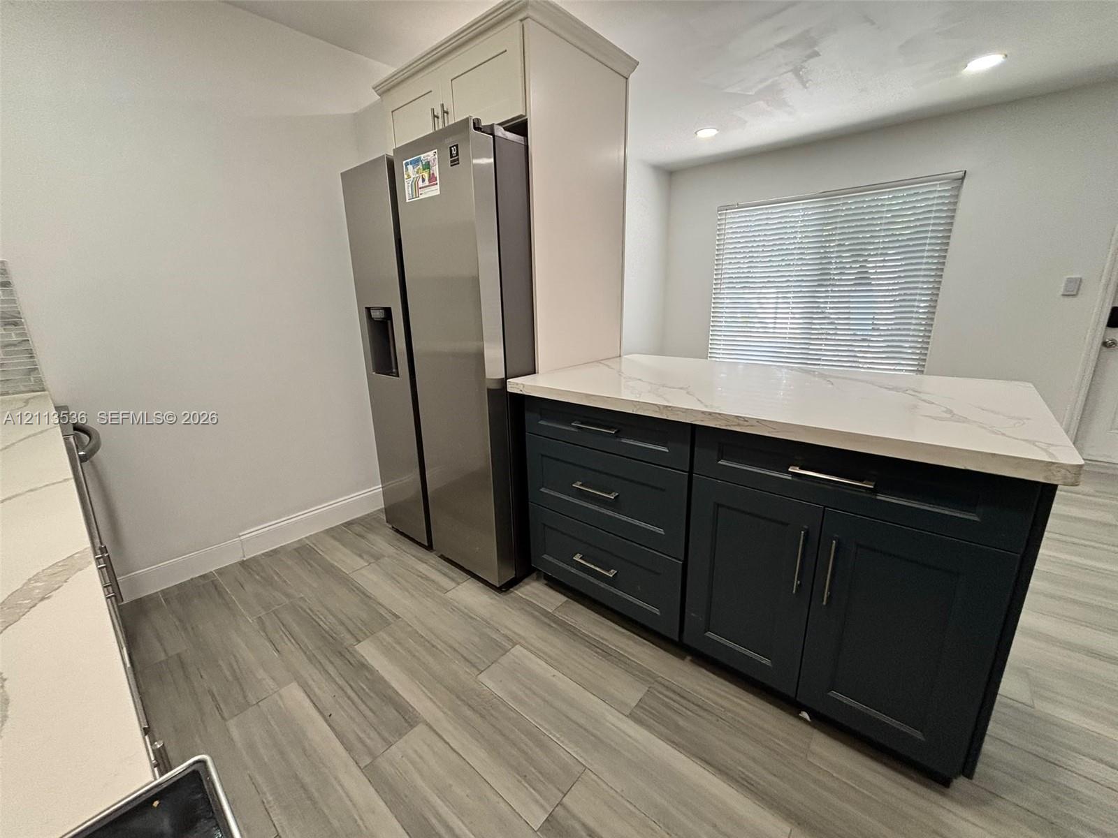 Kitchen featuring dark-toned cabinetry, light-colored countertops with veining, stainless steel refrigerator, wood-finish flooring, and recessed ceiling lighting Kitchen featuring dark-toned cabinetry, light-colored countertops with veining, stainless steel refrigerator, wood-finish flooring, and recessed ceiling lighting