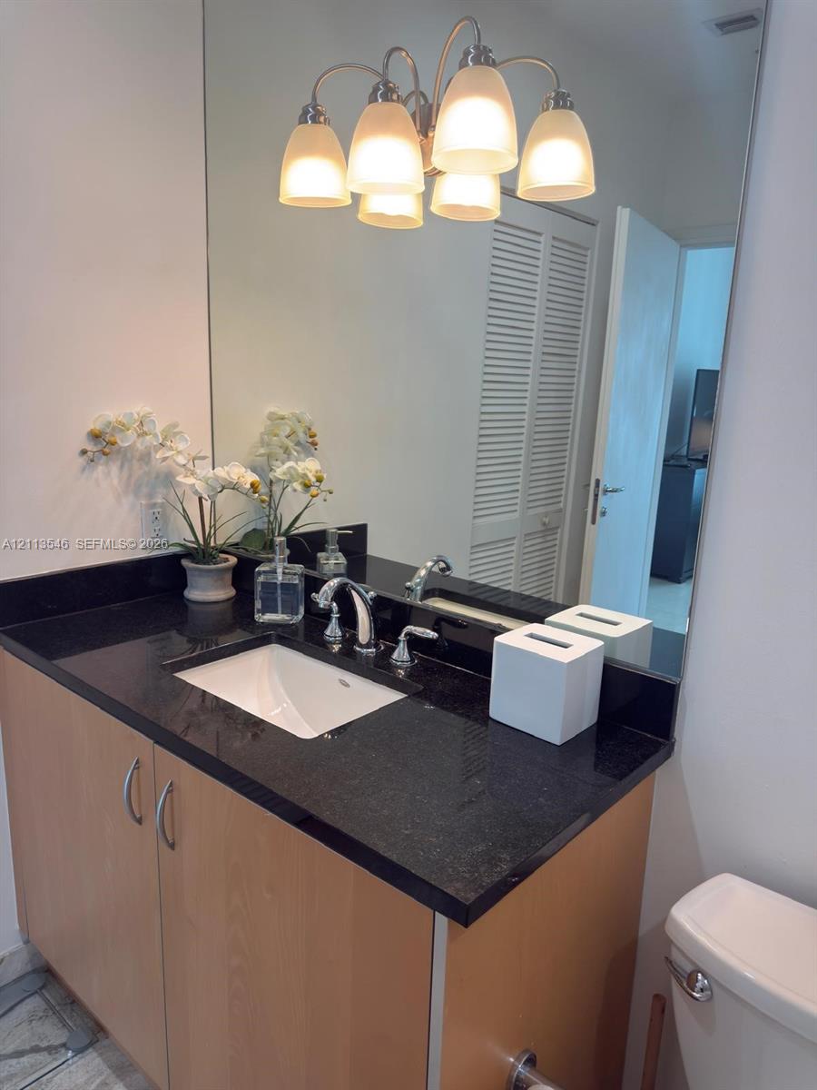 Bathroom vanity featuring a dark stone countertop, integrated rectangular sink, light wood-finish cabinetry with metallic pulls, a full-height wall mirror, and a multi-bulb light fixture
